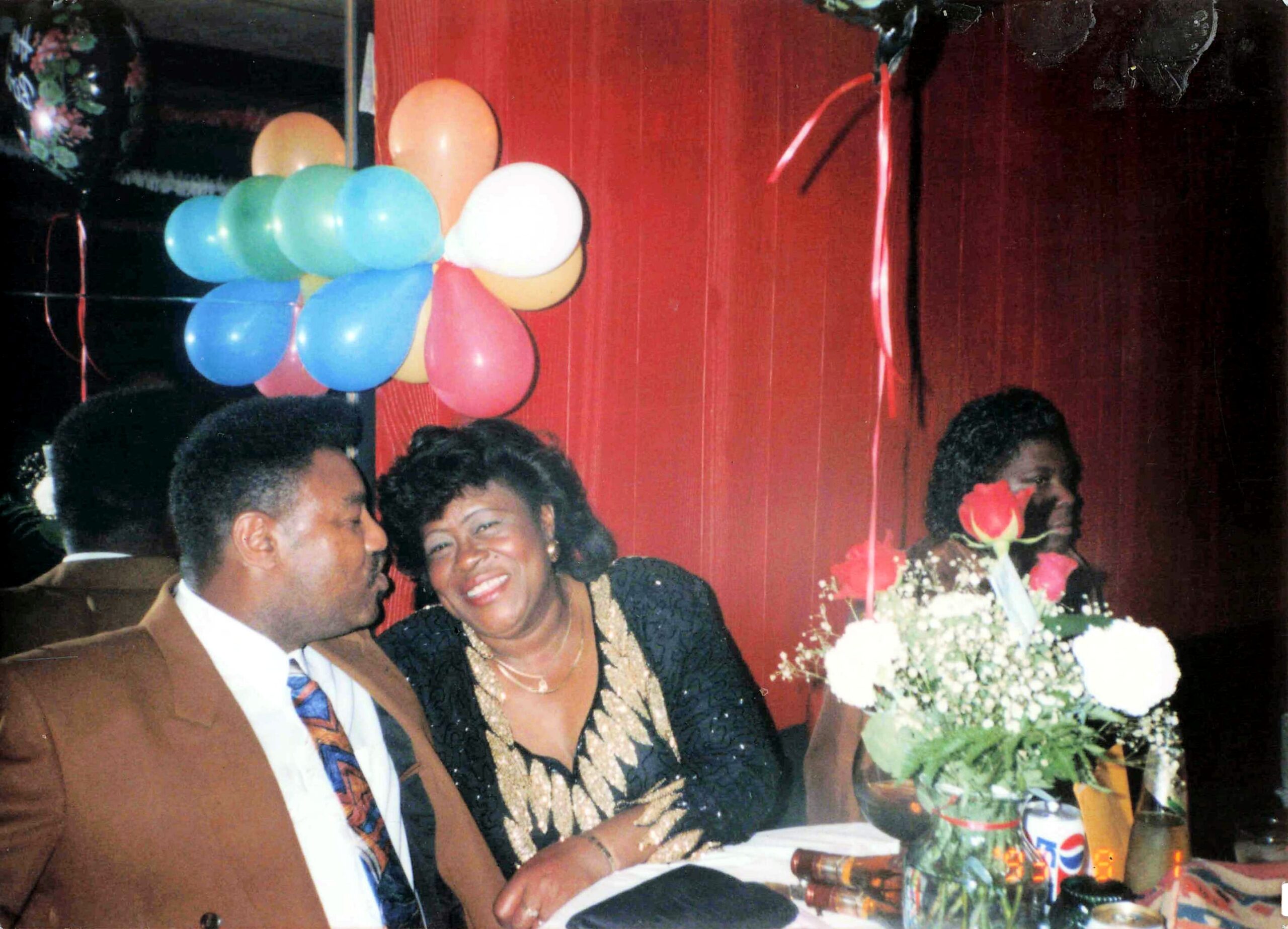 Smiling couple at a celebration table with colorful balloons and a red backdrop behind them.