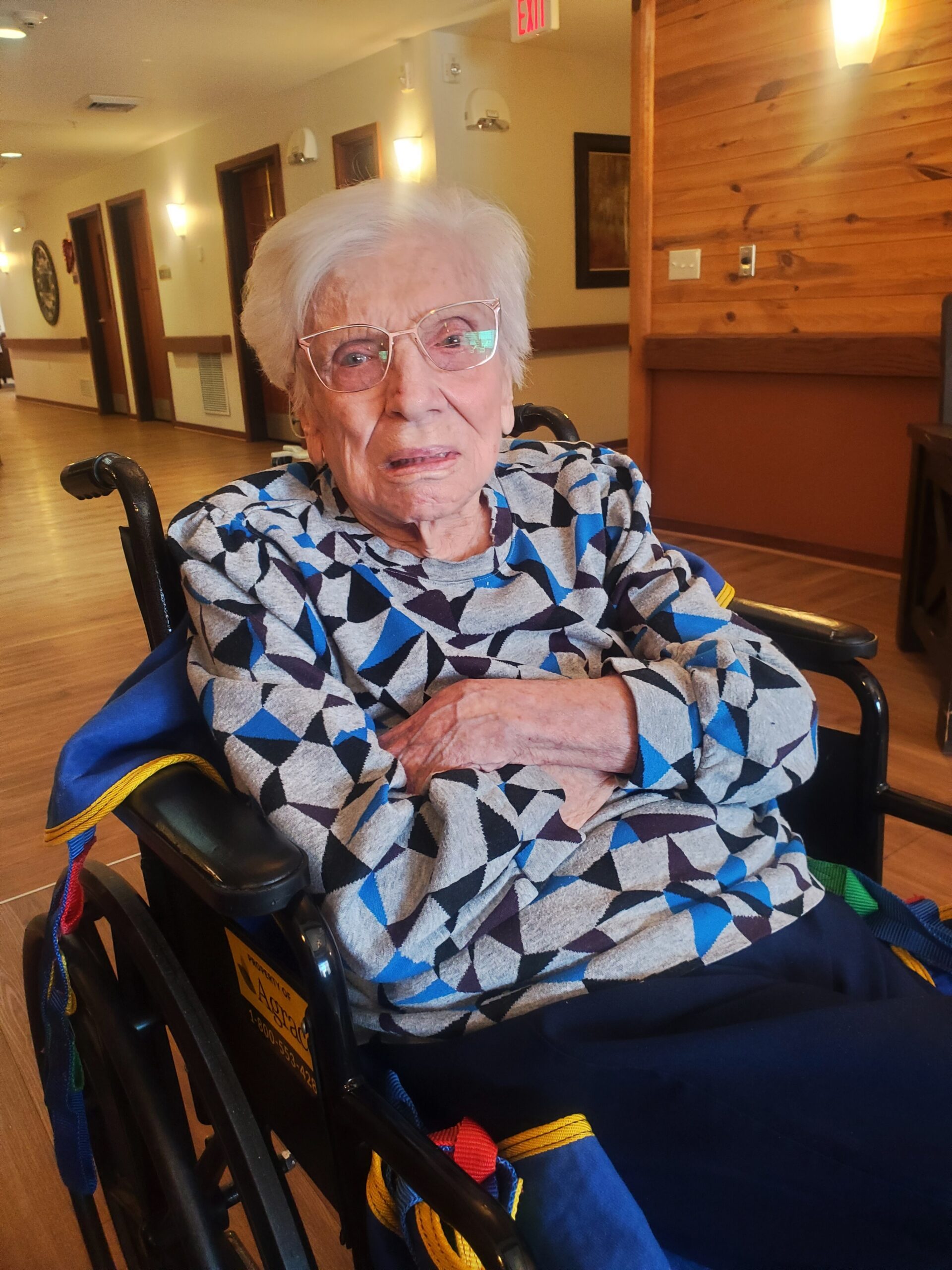 Older woman with white hair and glasses sitting in a wheelchair in a hallway of a care facility, wearing a blue, gray, and black geometric patterned top.