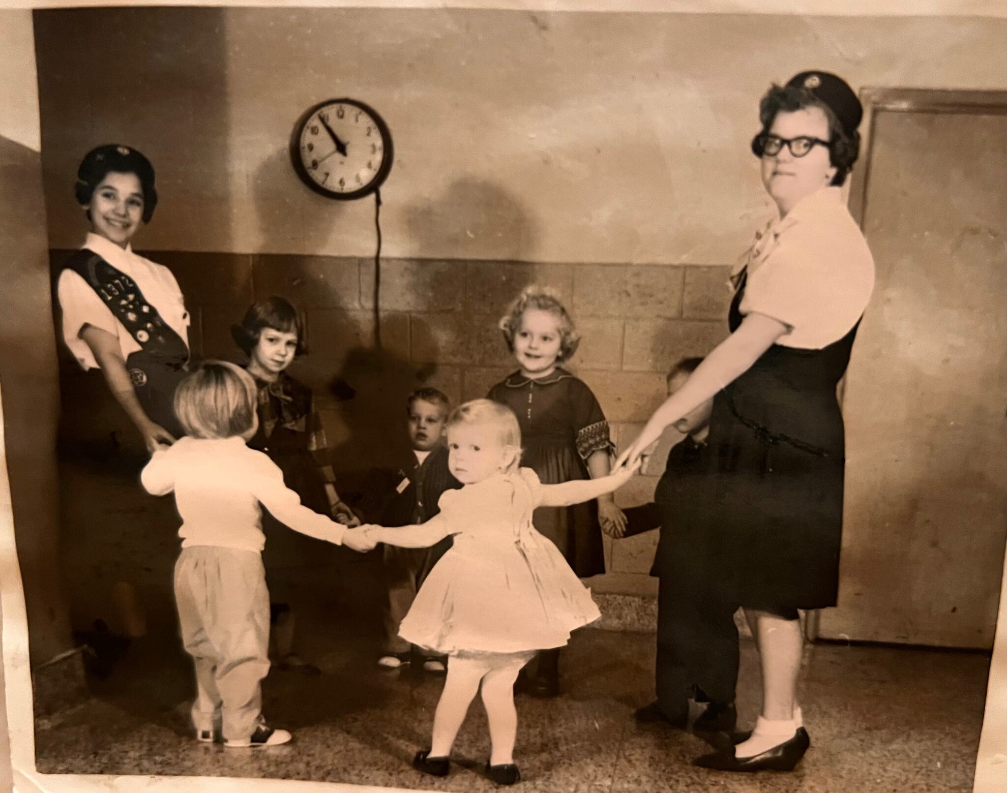 Children in dresses and tights hold hands in a circle, supervised by two women in vintage attire, with a clock on the wall behind them.