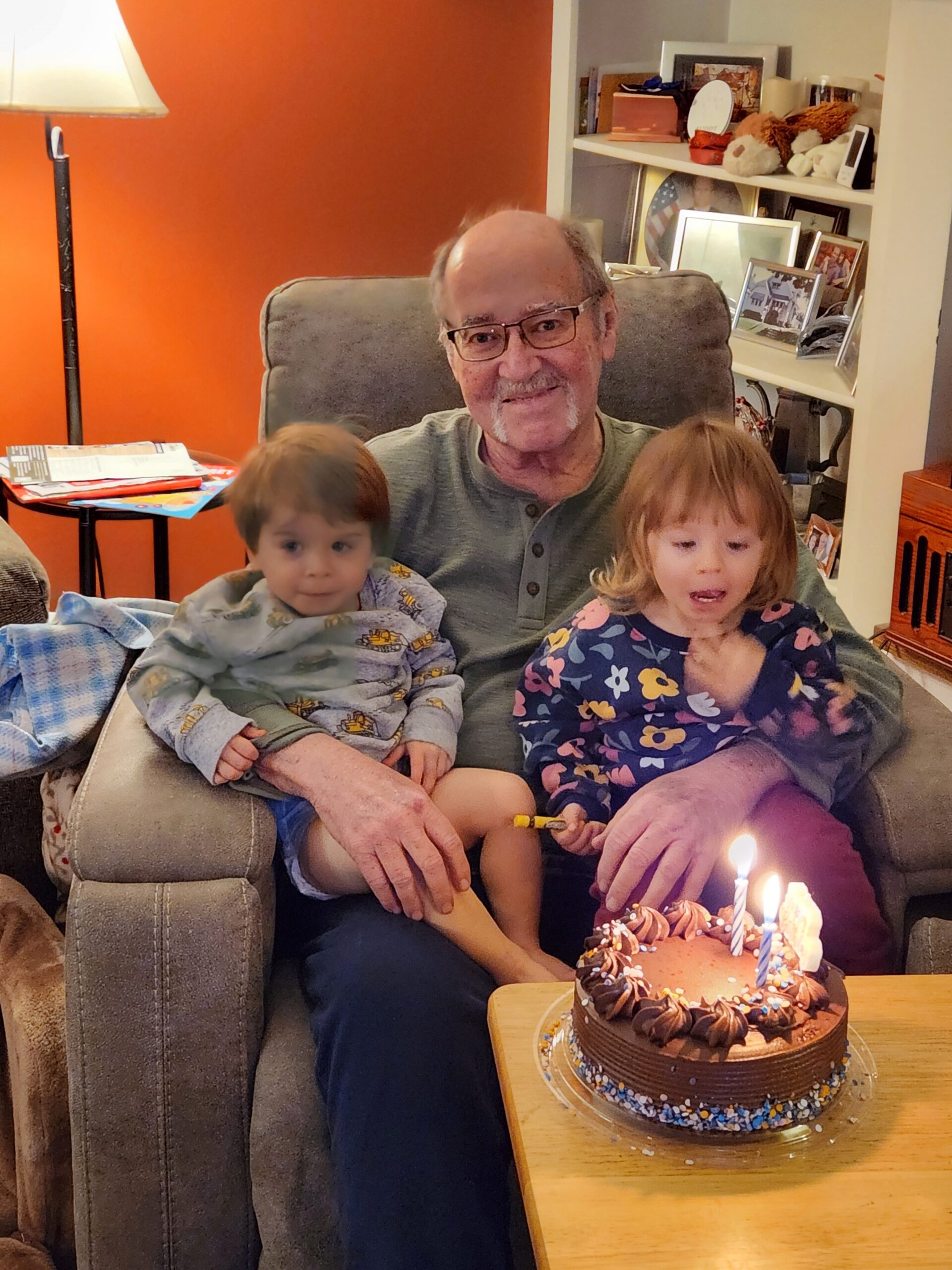 Grandfather with two young children on a couch during a birthday cake lighting moment, indoors by an orange wall.