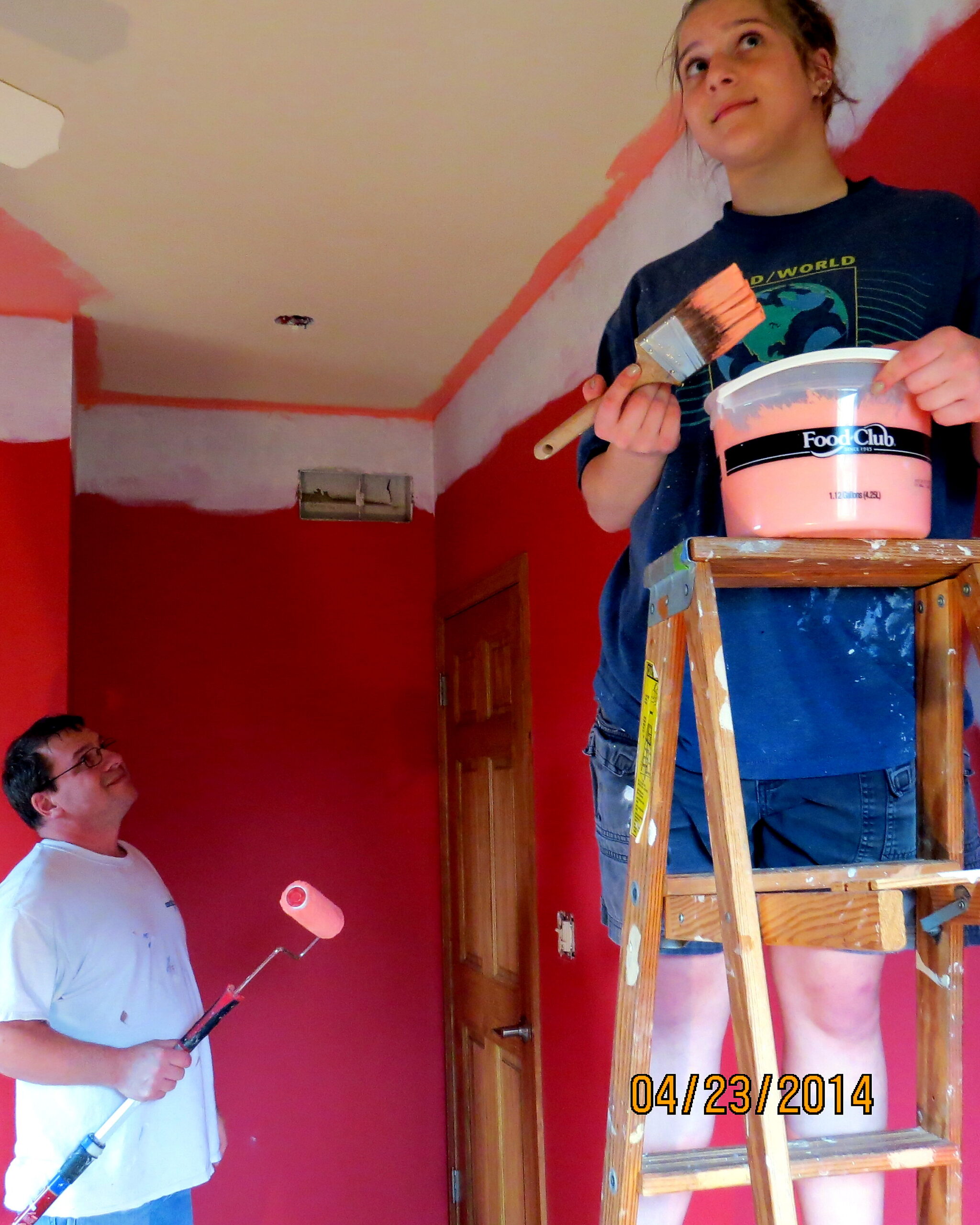 Person on a ladder holding a paint bucket and brush, painting a room with red walls; another man stands below with a paint roller to help.