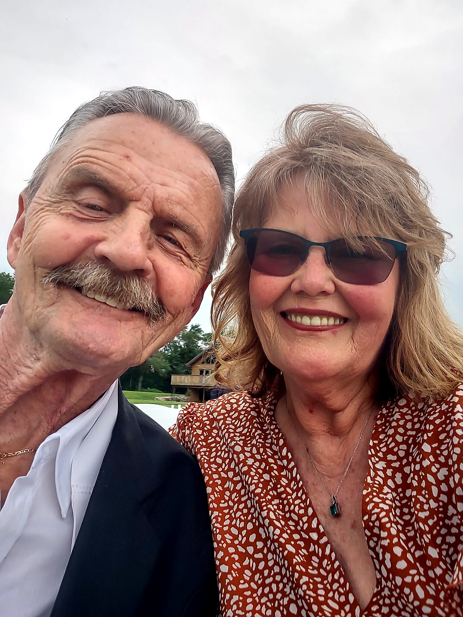Close-up of smiling older couple taking a selfie outdoors, man on left with suit jacket and white shirt, woman on right wearing sunglasses and a patterned blouse