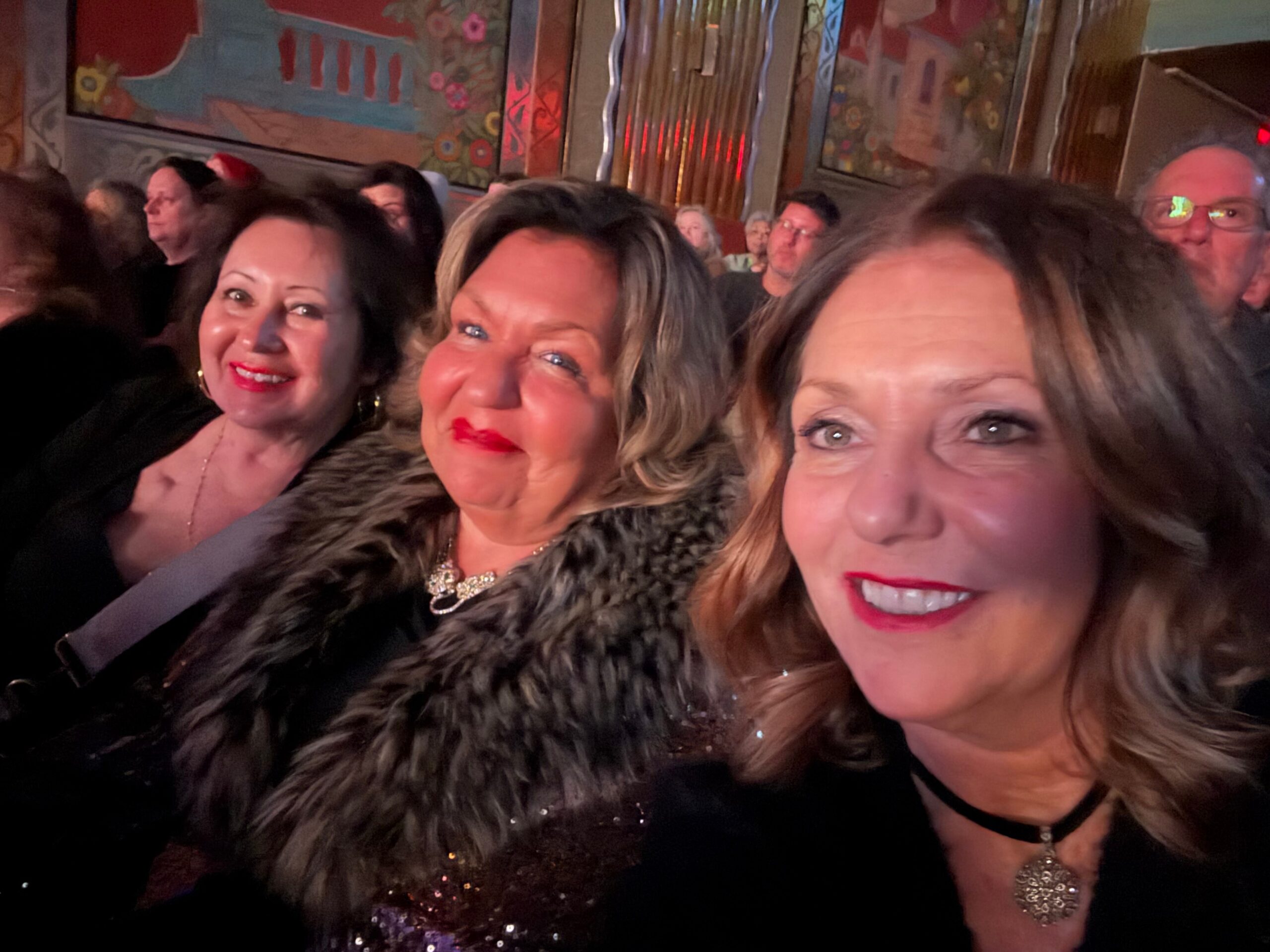 Close-up selfie of smiling women in a crowded party venue with a colorful mural in the background?