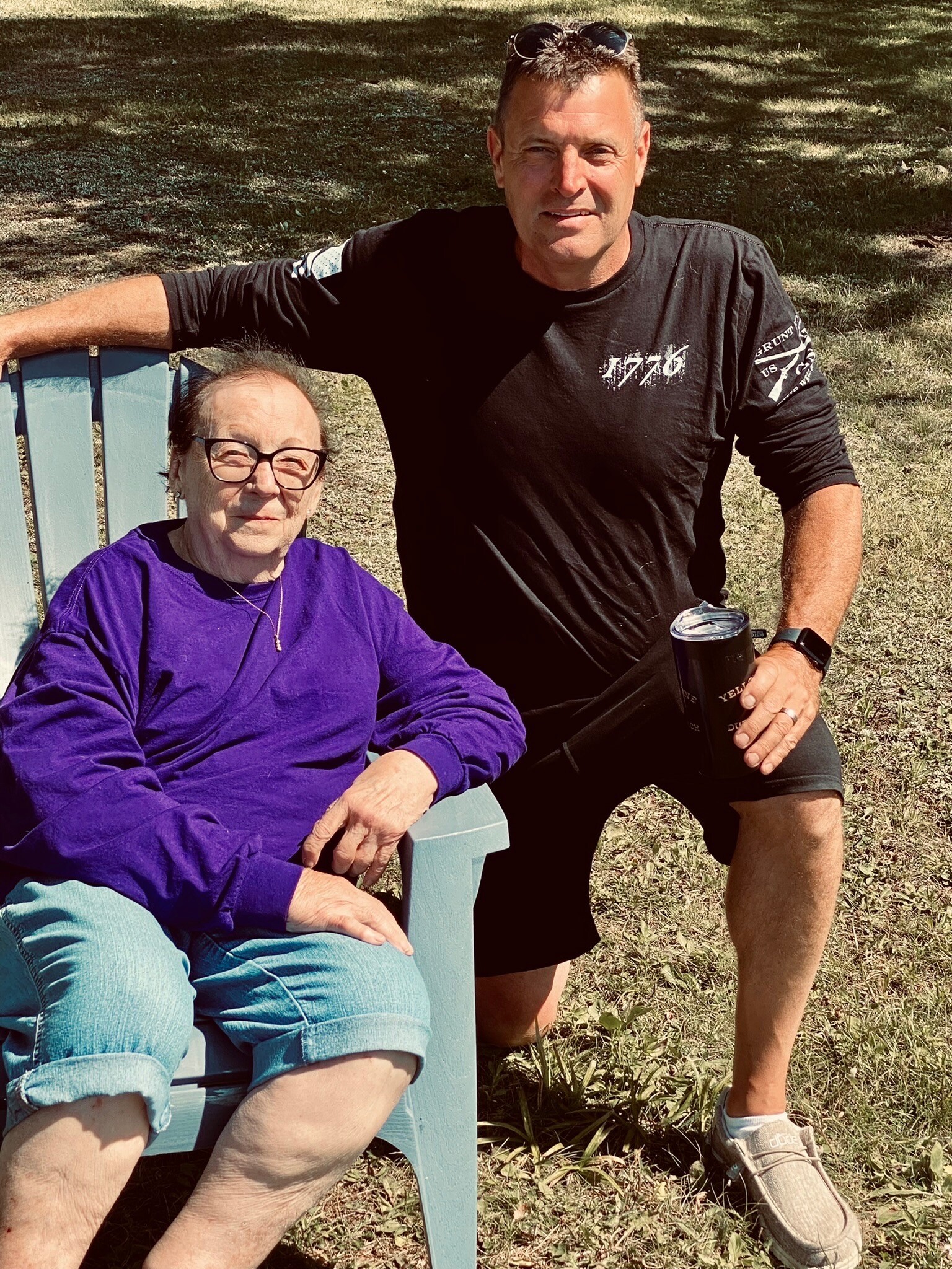 Older woman in a purple top and light jeans sits in a blue outdoor chair while a man in a black shirt stands beside her, resting his arm on the chair on a sunny day in a park.