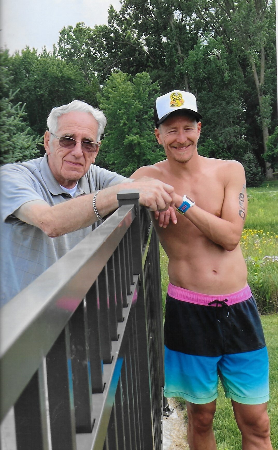 Two men lean on a black metal railing in a park, smiling at the camera; one older with glasses, the other shirtless in a white cap and colorful swim shorts.