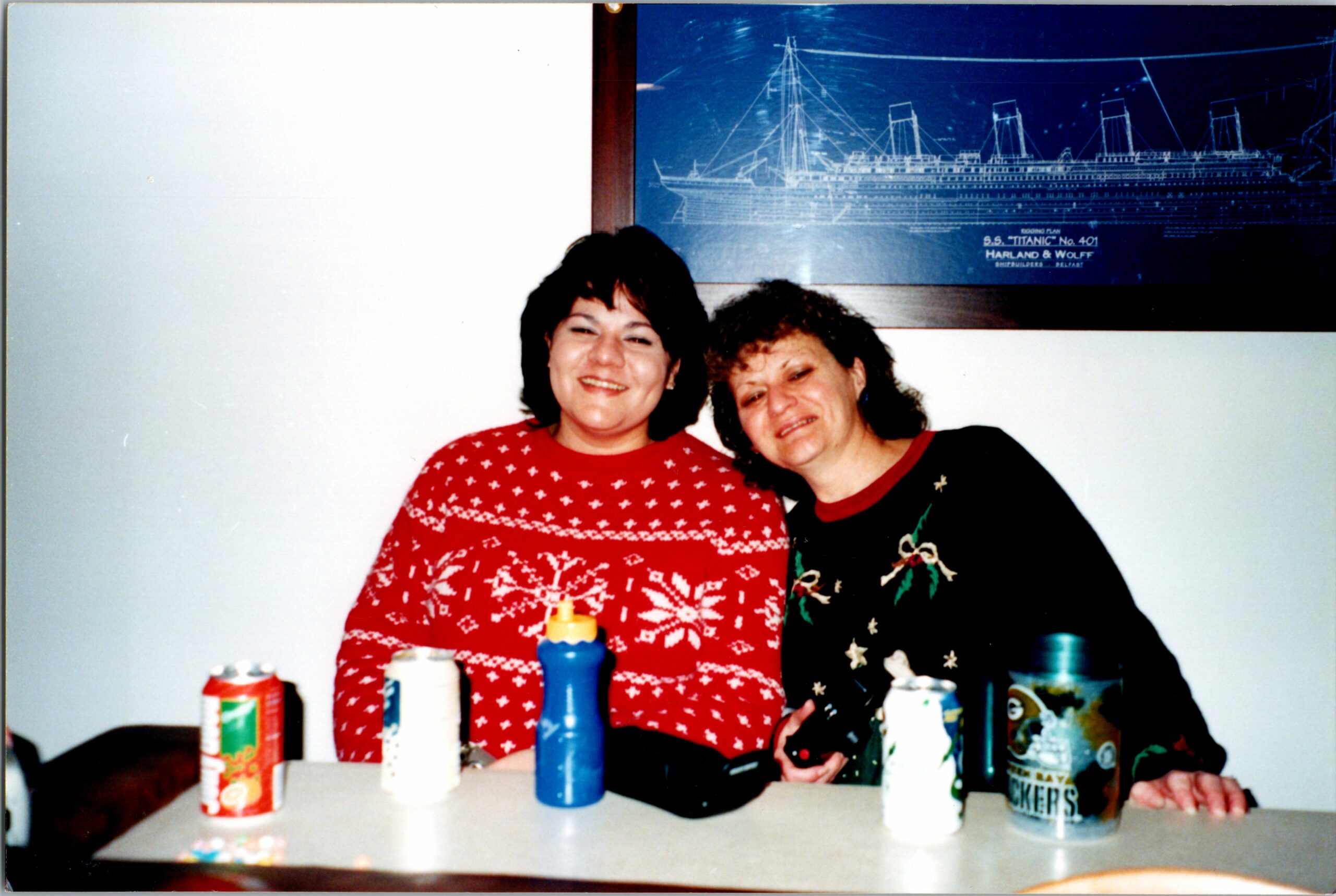 Two women in festive sweaters sit together at a table with drinks, smiling at the camera; a blue Titanic blueprint hangs on the wall behind them.
