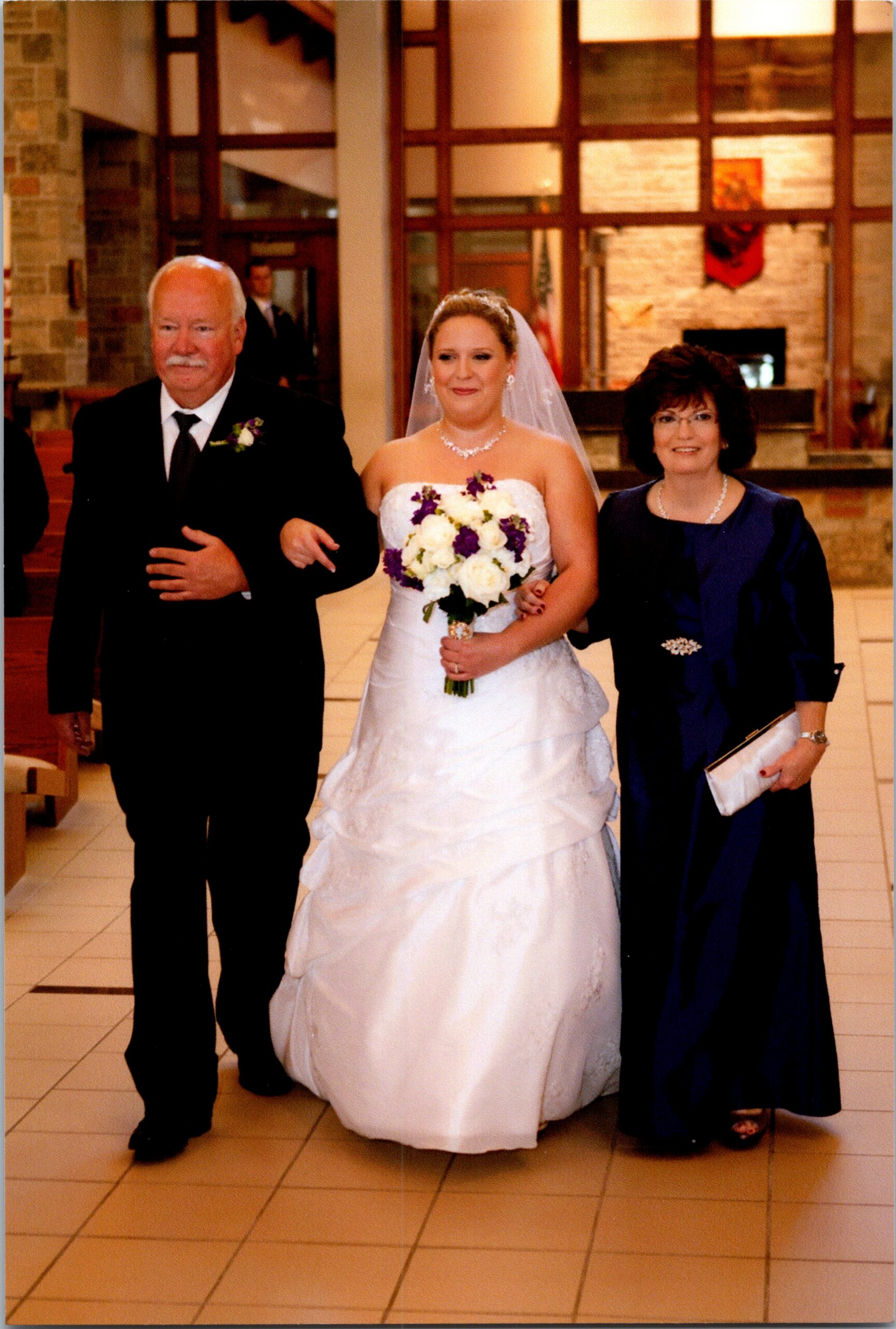Bride in a white wedding gown walks down the aisle flanked by her father in a tuxedo and her mother in a navy dress.