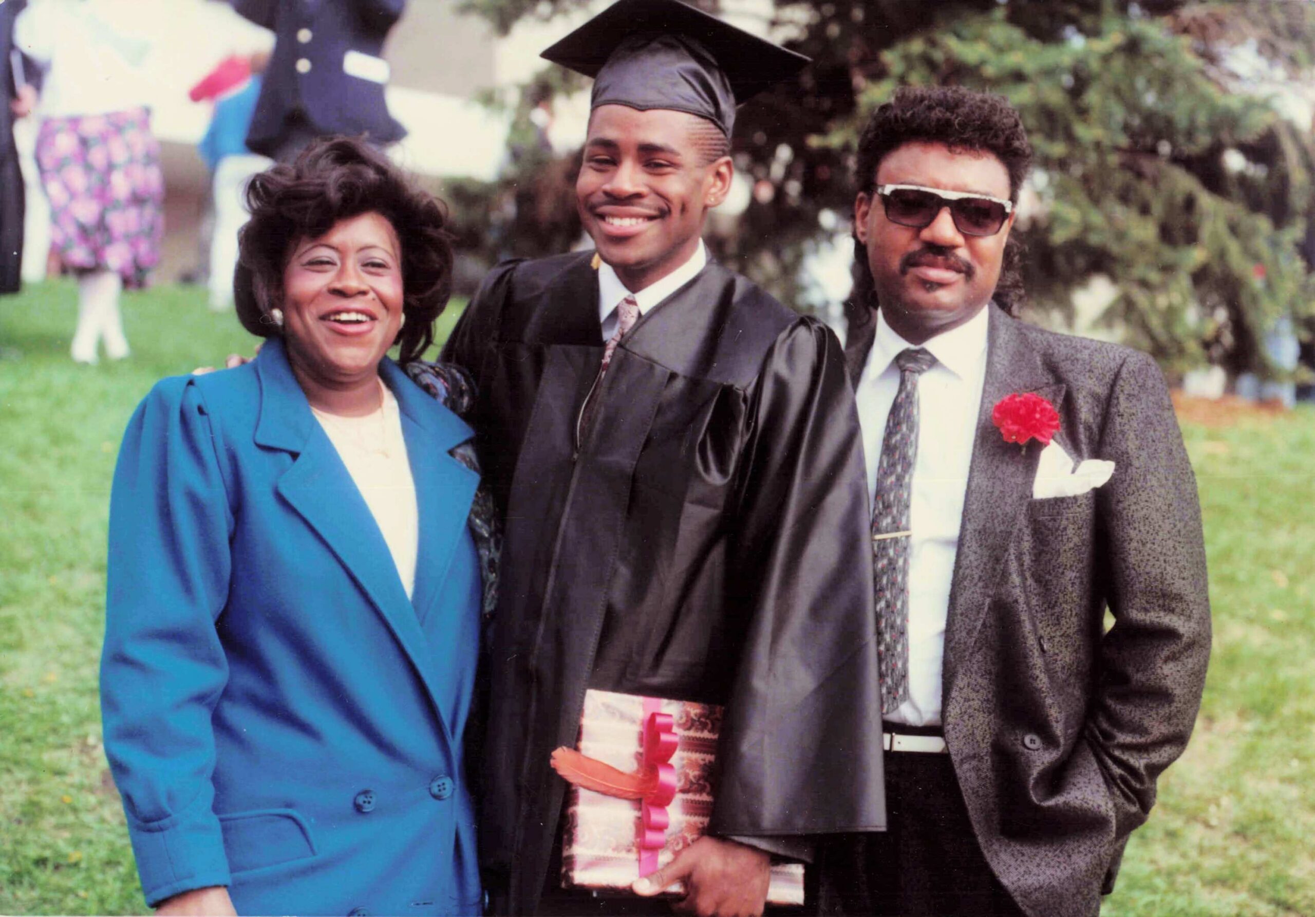 Graduate in cap and gown posing with two friends outdoors, smiling for a photo.