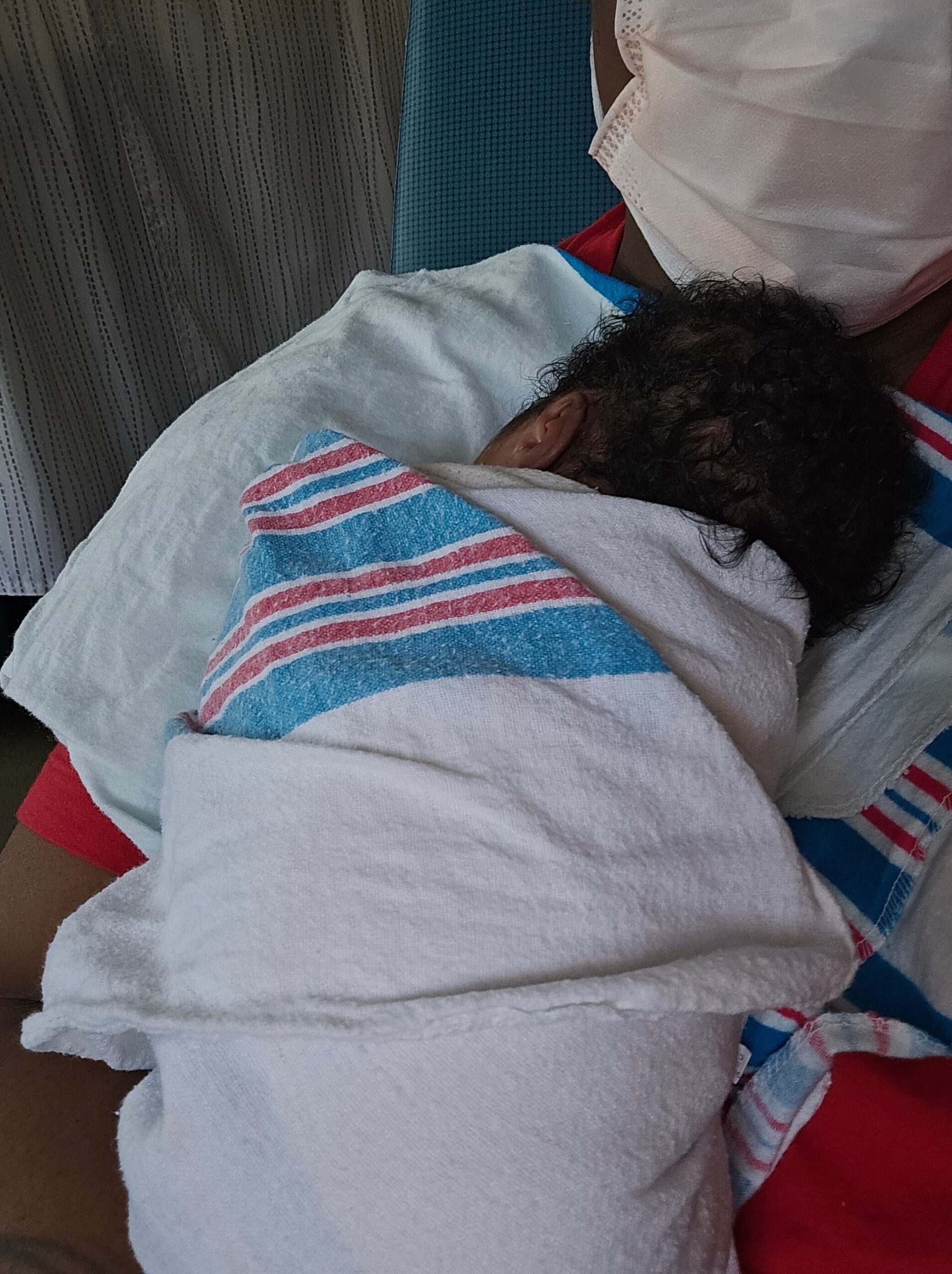 Newborn baby swaddled in a white towel with a blue and red striped cloth, resting on a caregiver's lap.