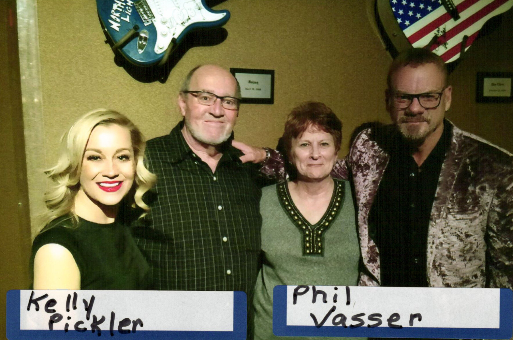 Four adults posing for a group photo backstage, with guitar decorations on the wall and an American flag in the background; signs identify Kelly Pickler and Phil Vasser.