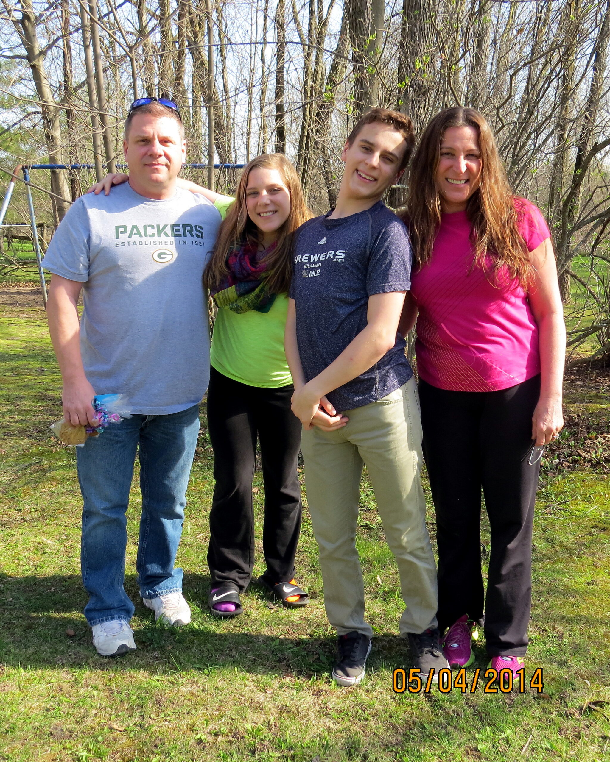 Four people stand arm-in-arm in a park, smiling at the camera: man in a gray Packers shirt, a girl in lime green, a young man in a navy shirt, and a woman in pink.