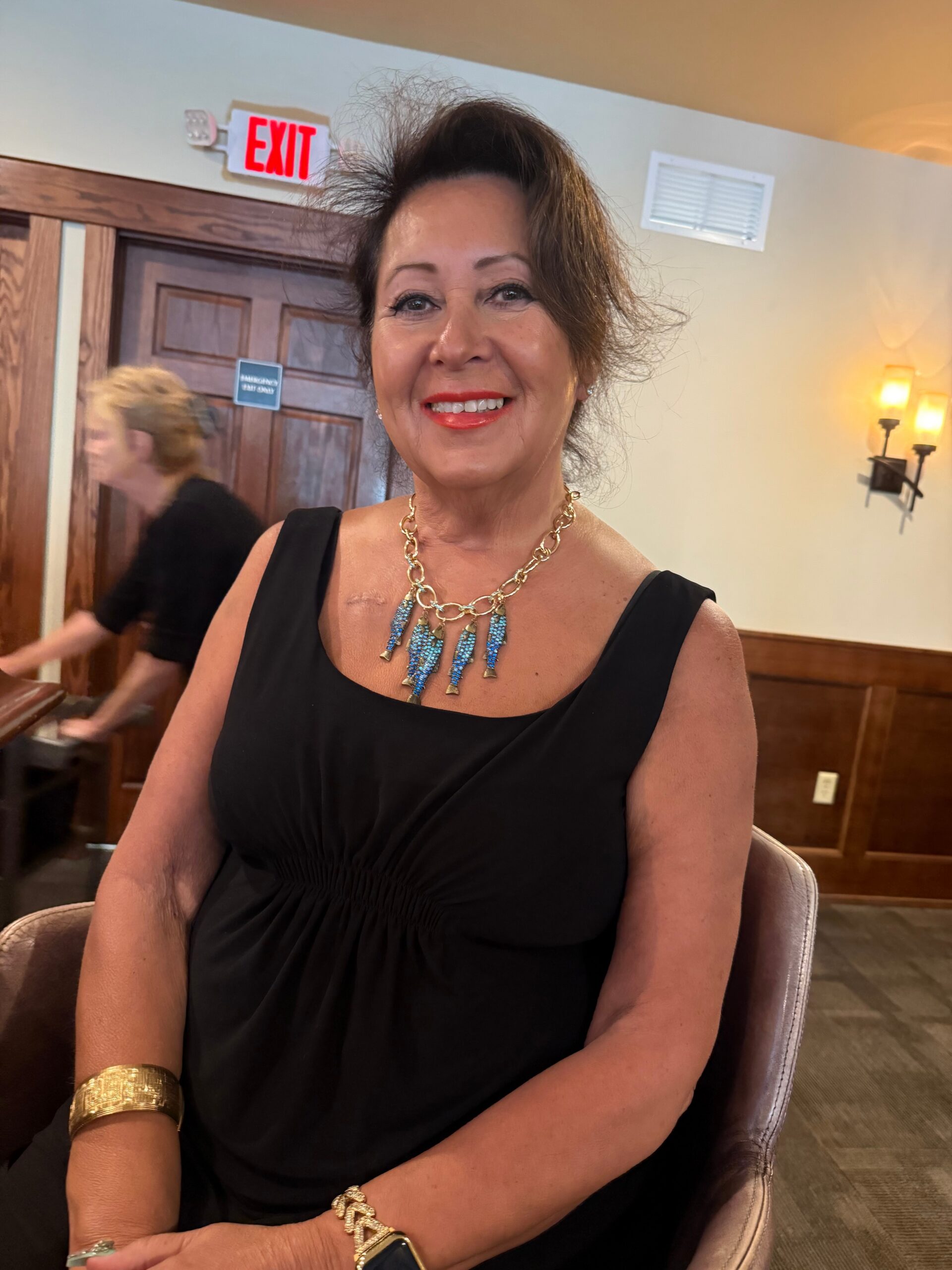 Smiling woman in a black sleeveless dress wearing a bold blue necklace and gold bracelets, indoors at a social event.