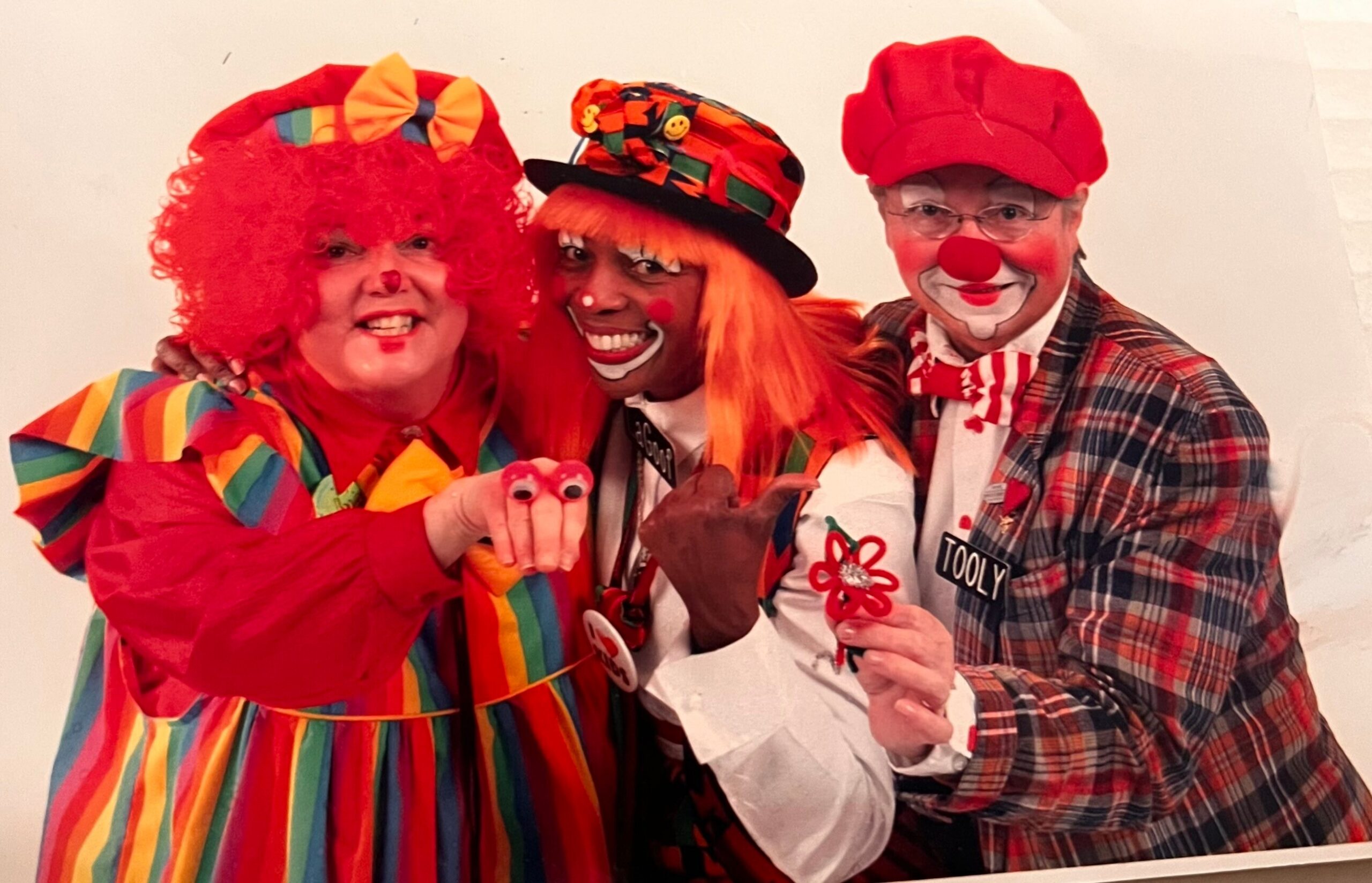 Three clowns posing together for a photo in bright, playful costumes: left in a red wig and rainbow-striped outfit, center with orange wig and hat, right wearing a red nose and plaid suit.