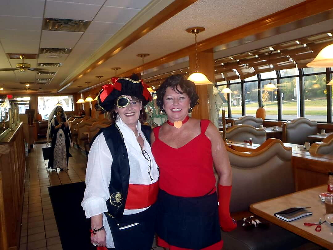 Two women in costumes posing together in a diner, one dressed as a pirate with hat and vest, the other in a red outfit with gloves and choker.