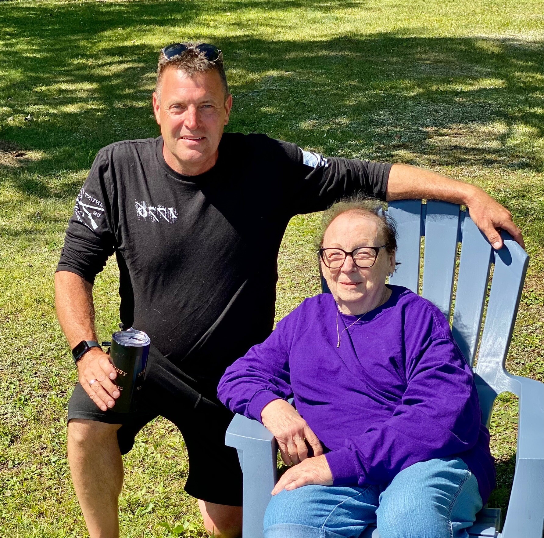 Man standing next to a seated older woman in a purple top on a sunny grassy day in a park.