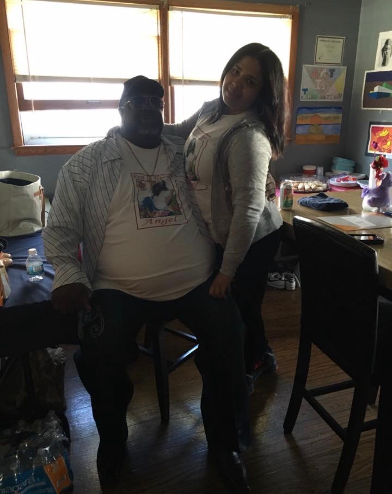 Two people pose indoors together: a man seated in a striped shirt and graphic tee, and a woman standing beside him, with art on the wall and snacks on a table in the background.