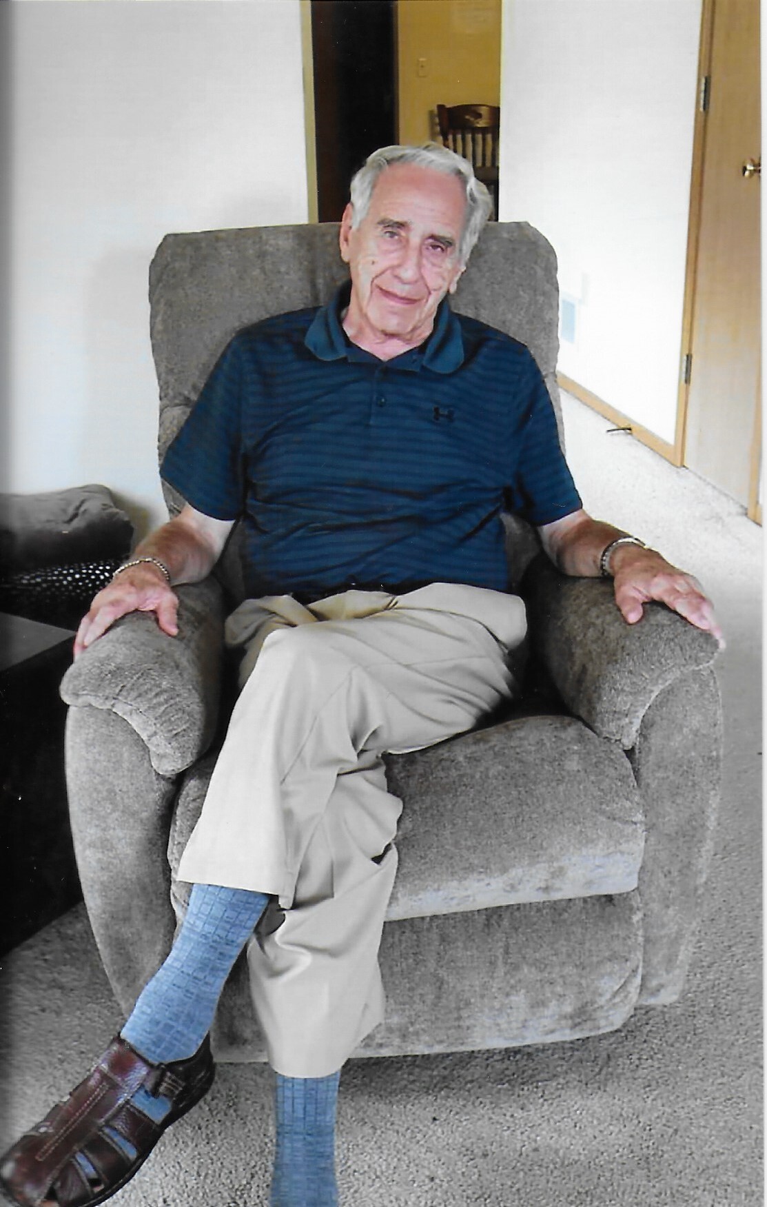 An elderly man with white hair sits in a gray upholstered armchair in a living room, wearing a blue polo shirt, khaki pants, blue socks, and brown shoes.