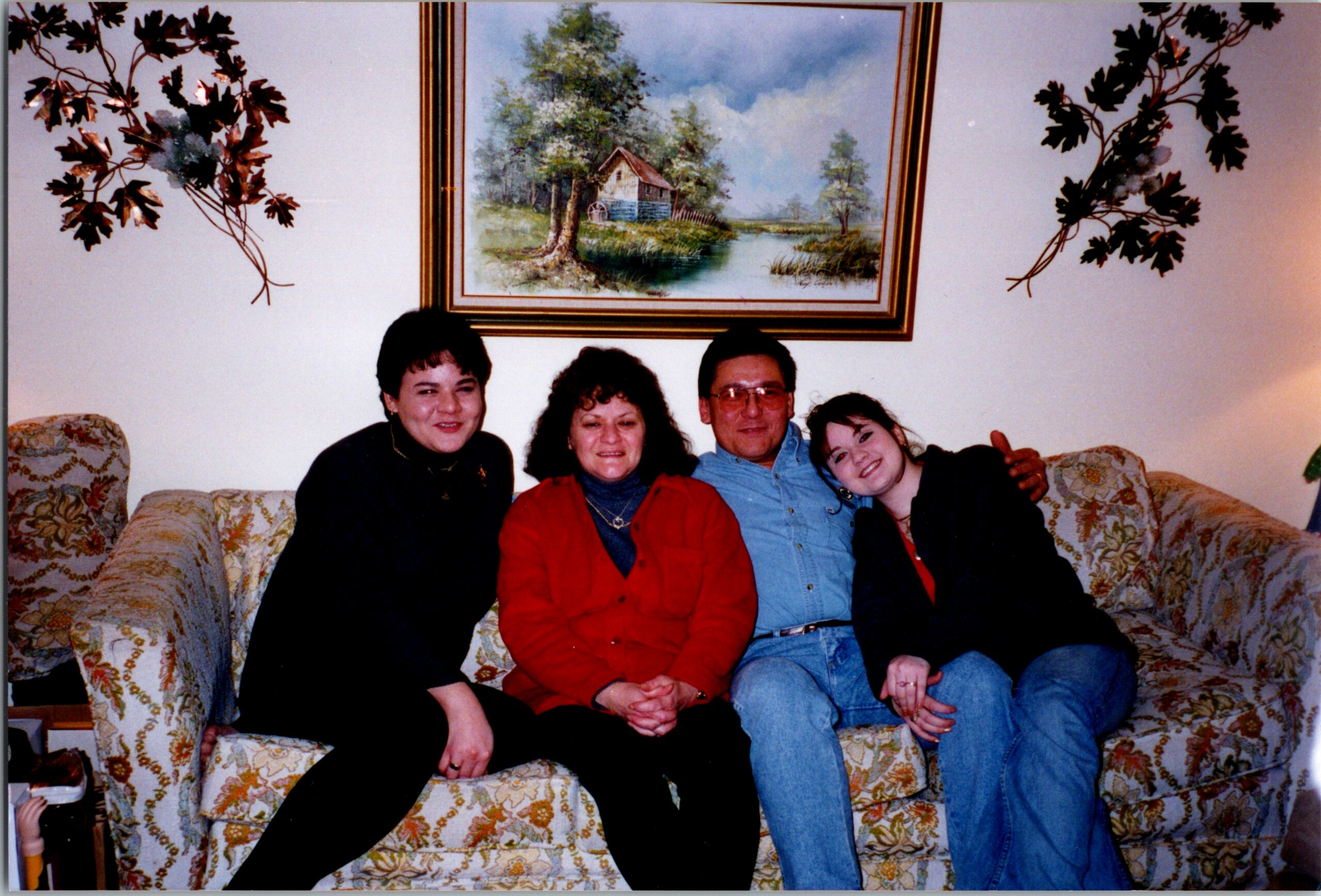 Four people sitting on a floral sofa in a living room, smiling; a framed landscape painting hangs on the wall behind them.