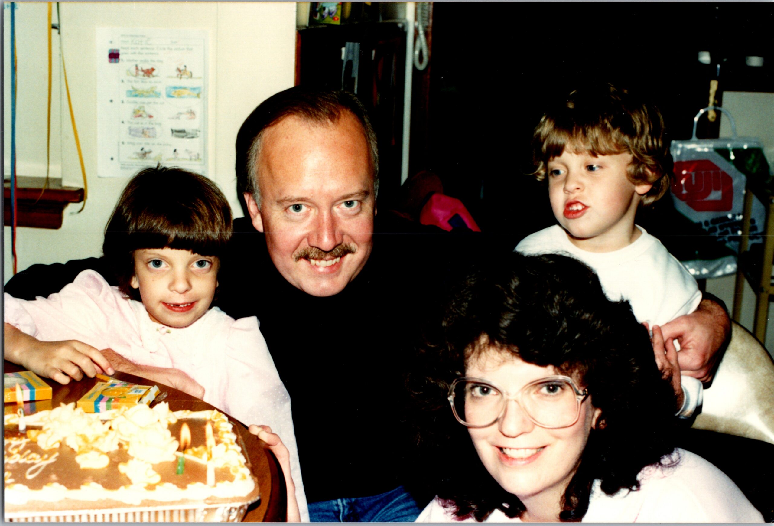 A man with a mustache and three children gathered around a birthday cake with frosting and candles.