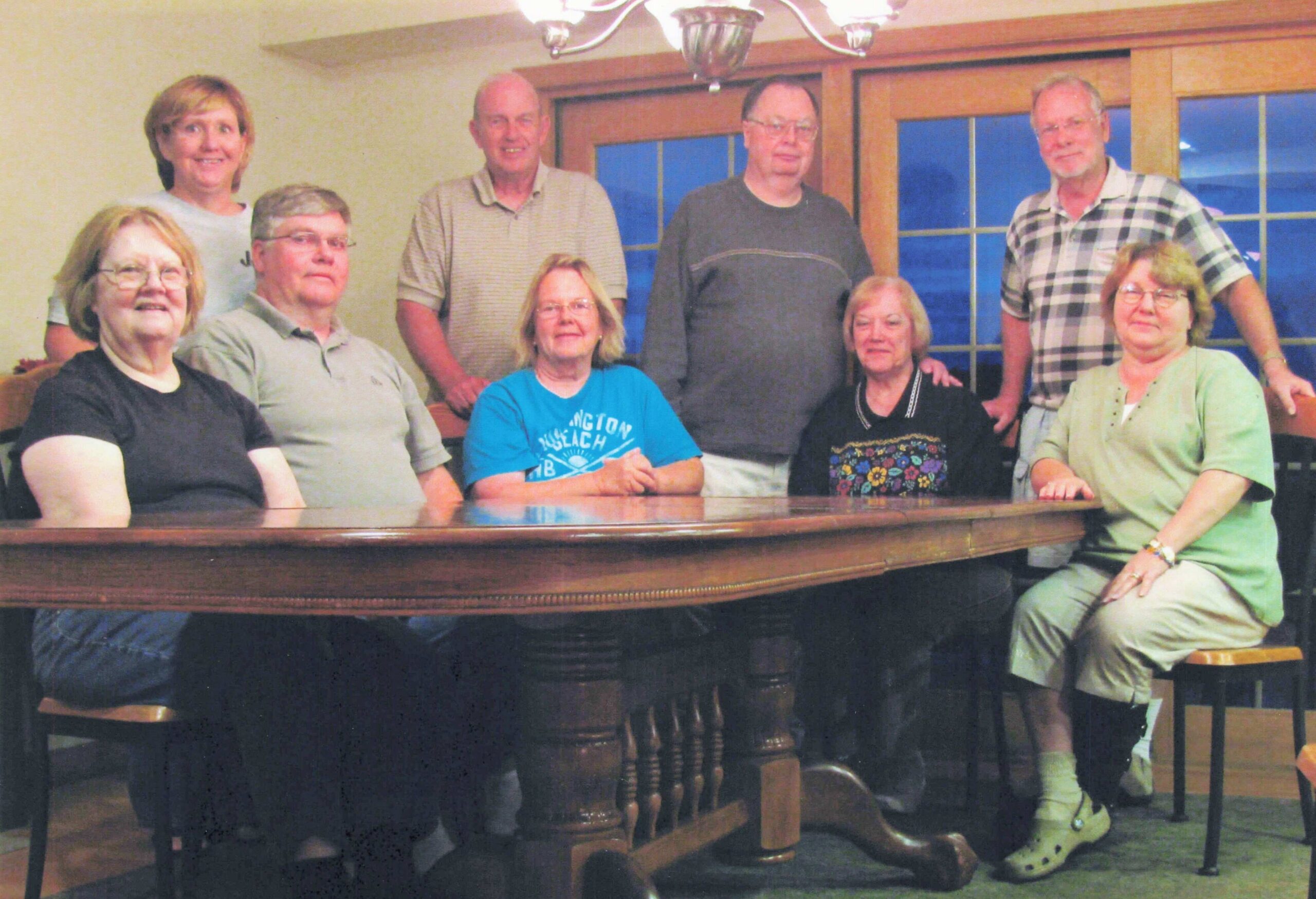 Eight adults pose around a polished piano in a casual indoor room, smiling at the camera.