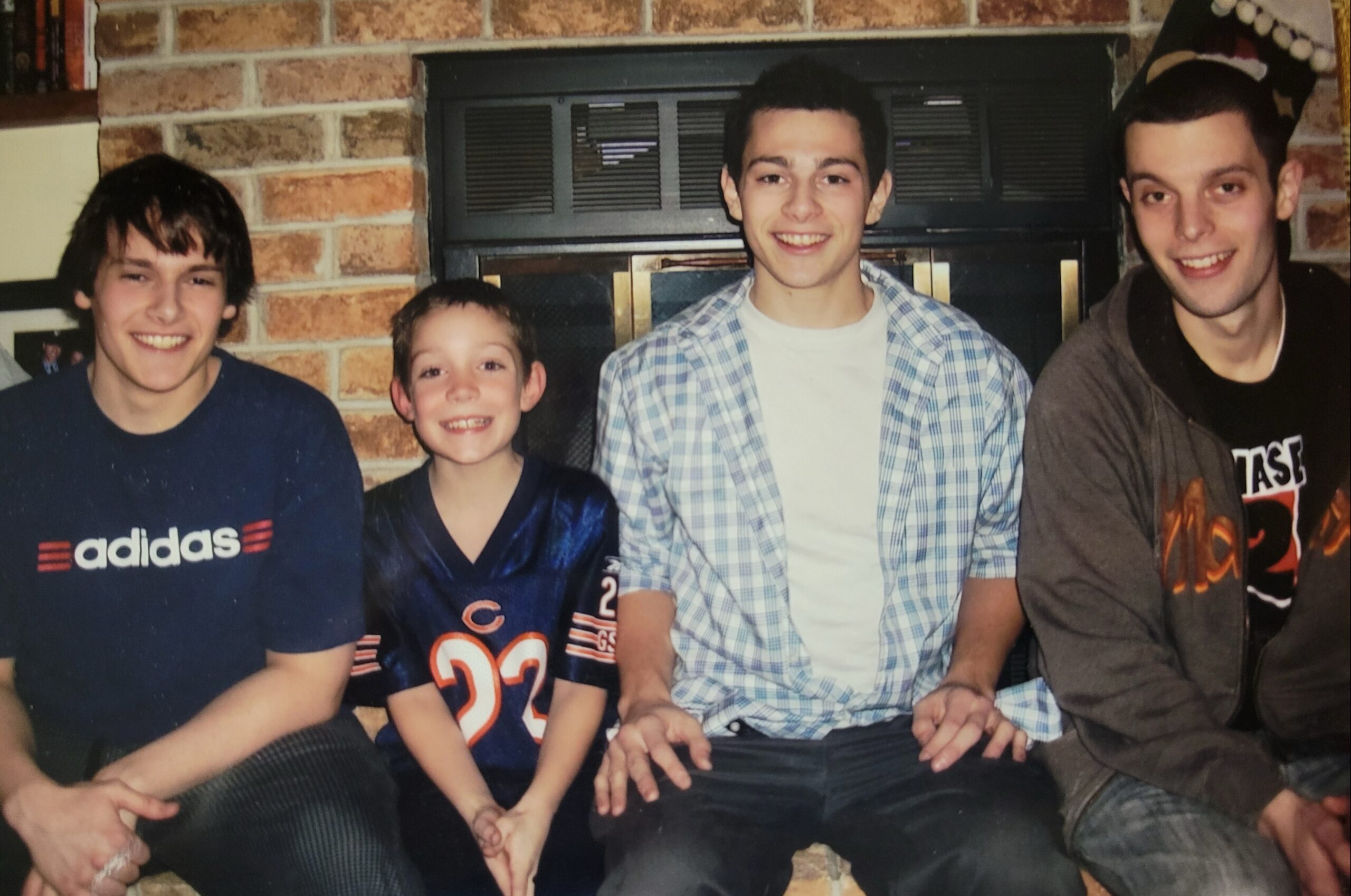 Four young men posing for a photo on a couch in front of a brick fireplace, all smiling at the camera with their hands resting on their laps.