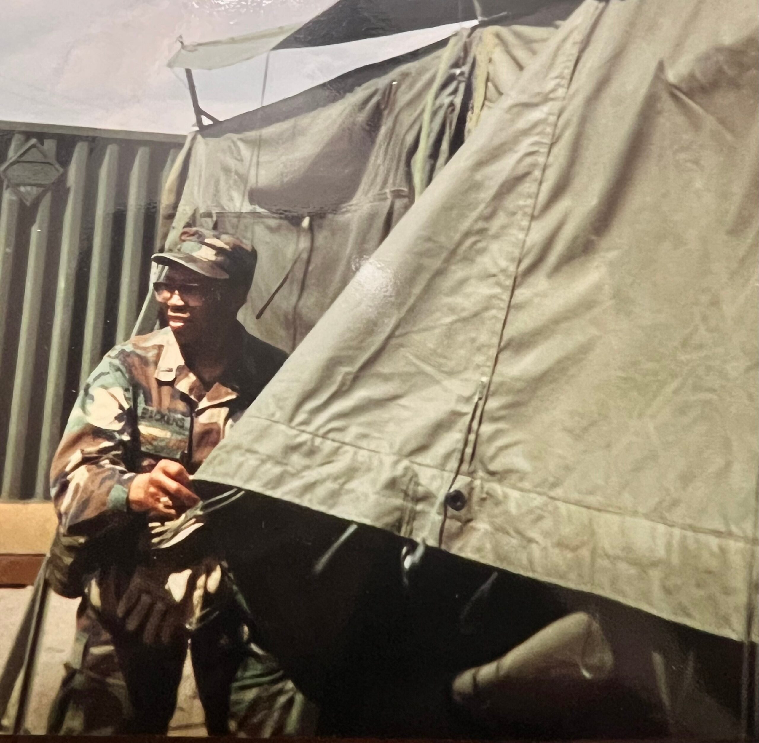 Soldier in camouflage uniform standing beside a tent-like beige fabric indoors, wearing a cap and looking to the side.
