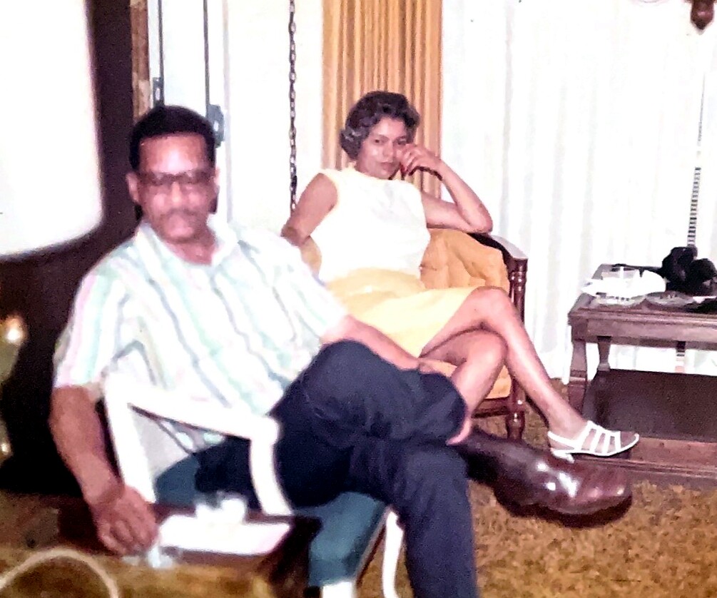 Man in striped shirt sits in a white plastic chair beside a woman in a yellow skirt and white top in a living room. A coffee table with a phone and a guitar is visible in the background.