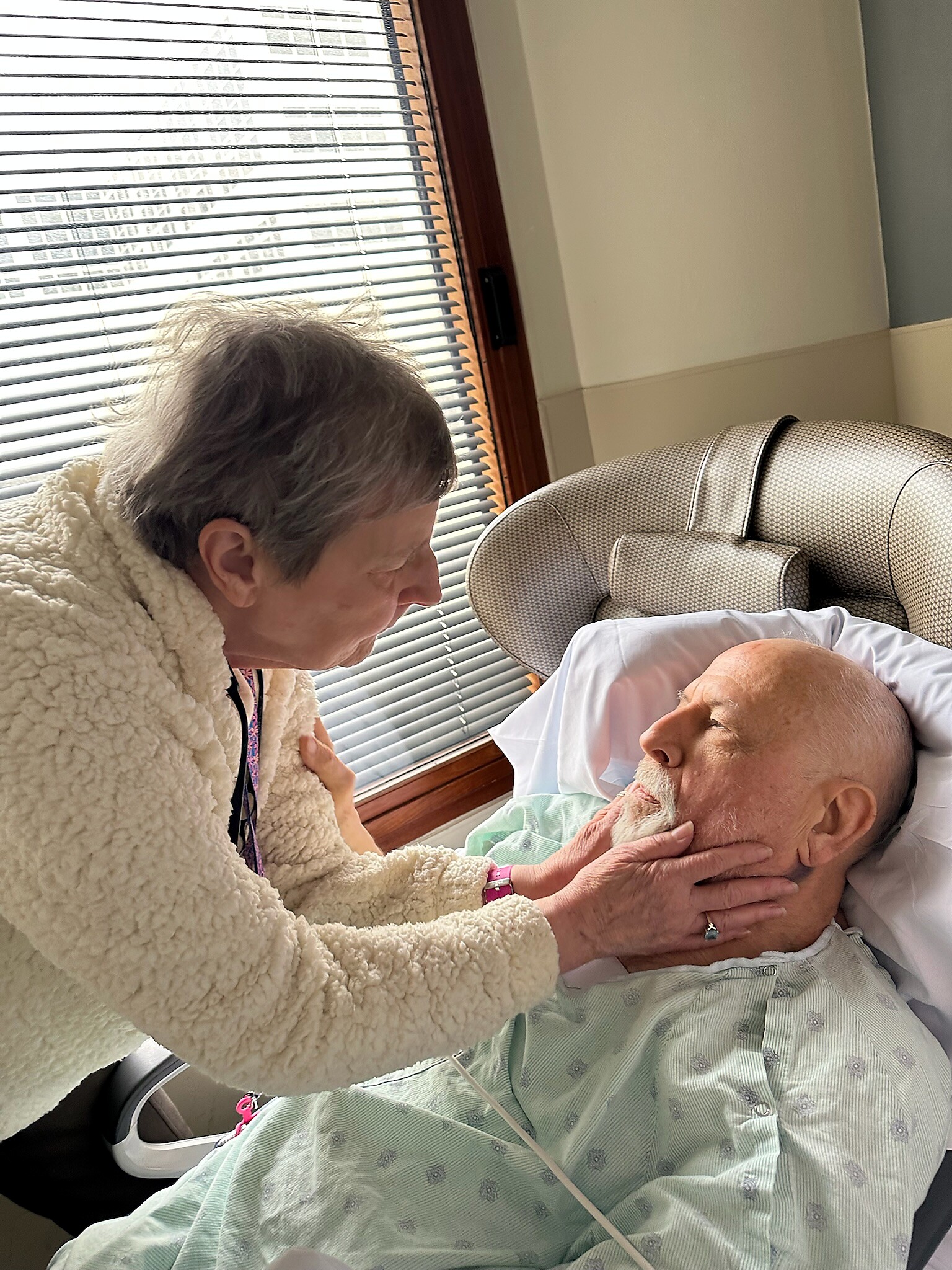 Caregiver gently holding an elderly man's face as he rests in a hospital bed by a window with blinds.
