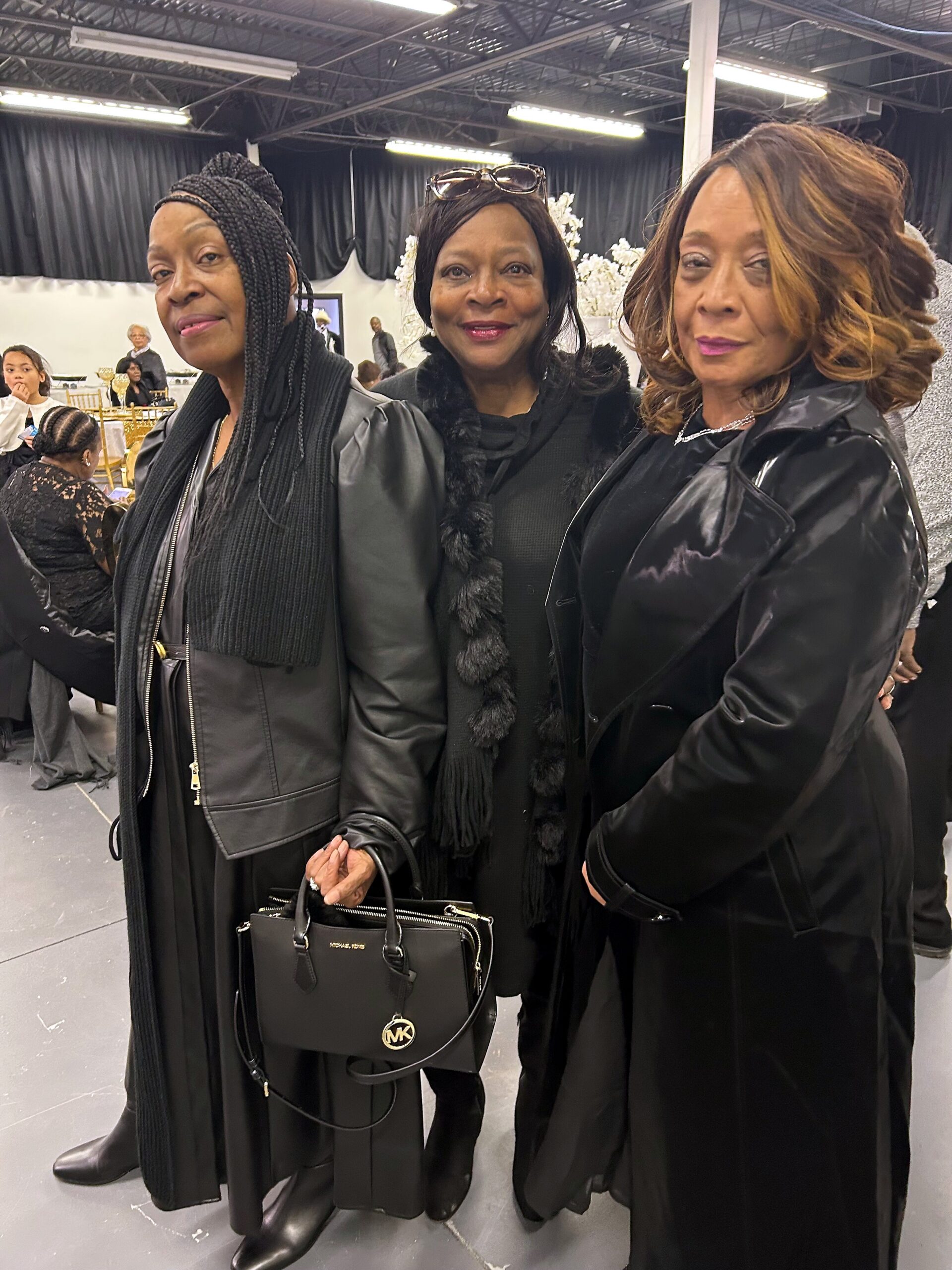 Three stylish women posing together indoors at a fashion event, all in black outfits and accessories.
