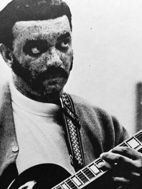 Black-and-white photo of a man with a mustache playing a guitar, looking to the side.