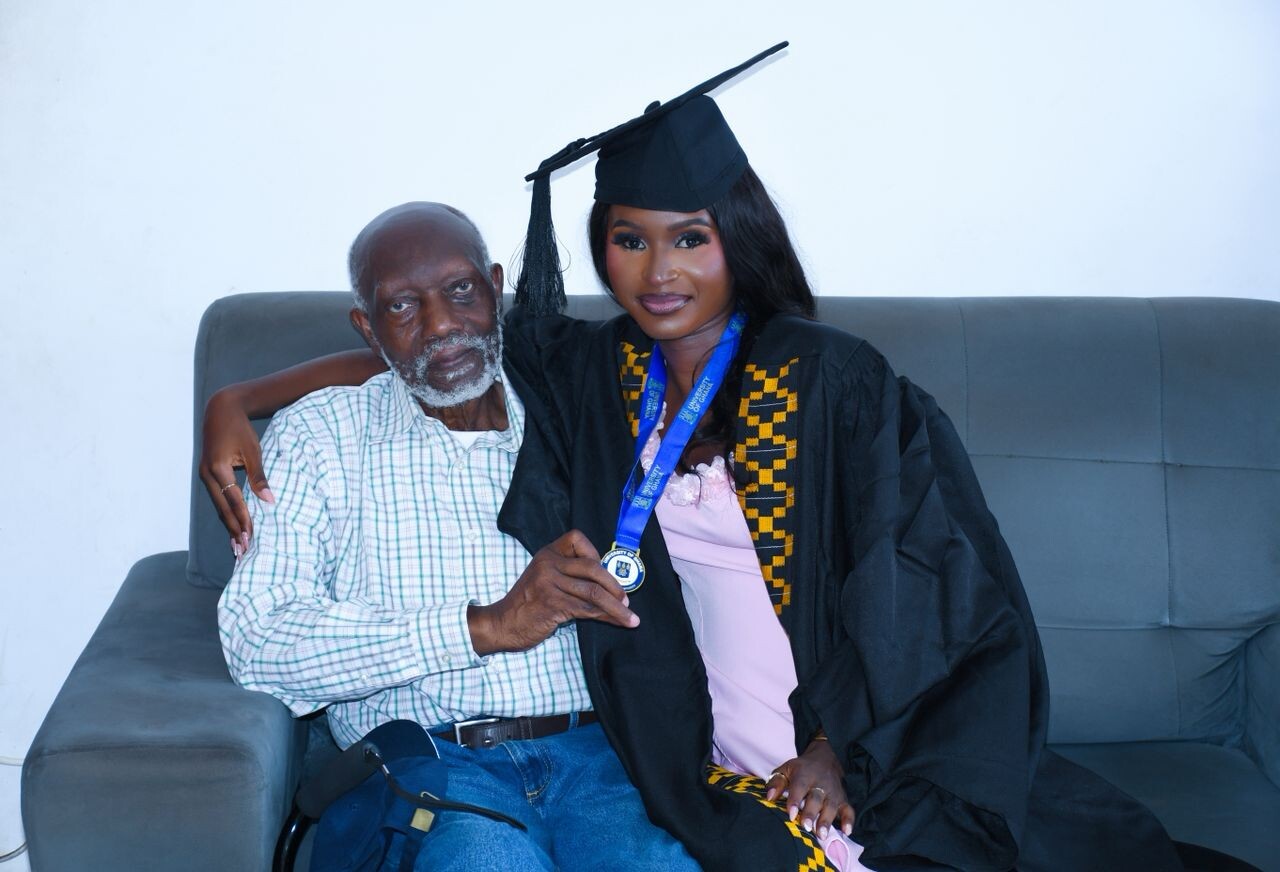 Graduate woman in cap and gown sits with a smiling older man on a sofa, medal visible around her neck.