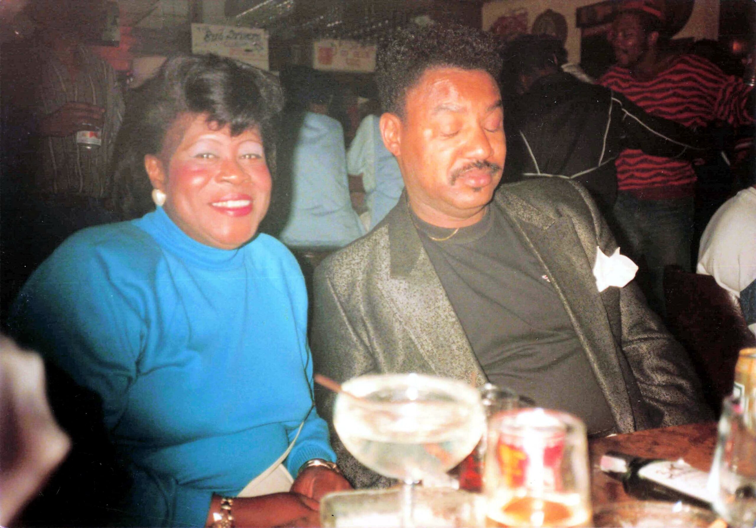 Two people sit at a table during a social gathering: a smiling woman in a bright blue top on the left and a man in a dark suit with eyes closed on the right, with drinks in front of them.