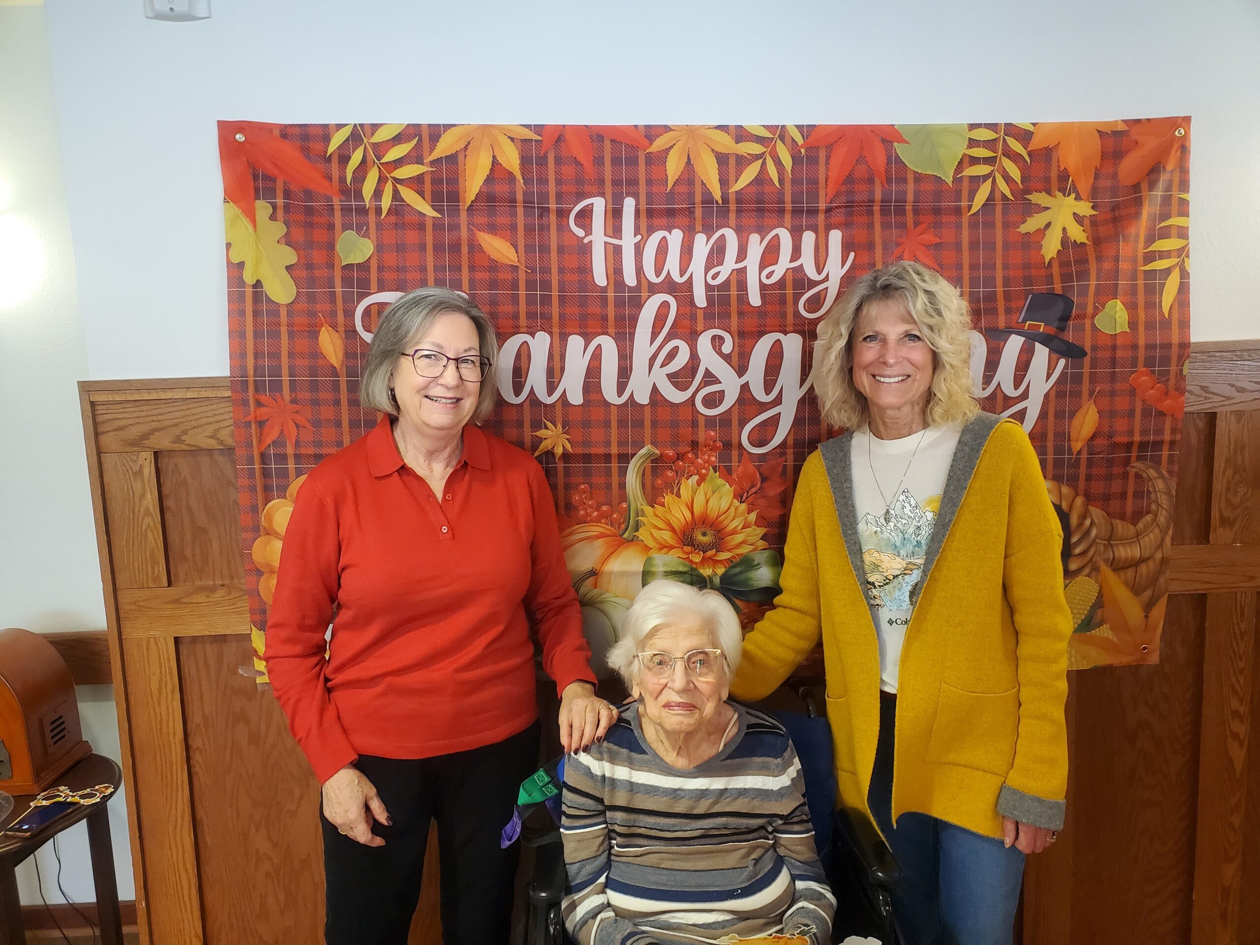 Three women stand around an elderly woman in a wheelchair, posing in front of a 'Happy Thanksgiving' autumn backdrop.