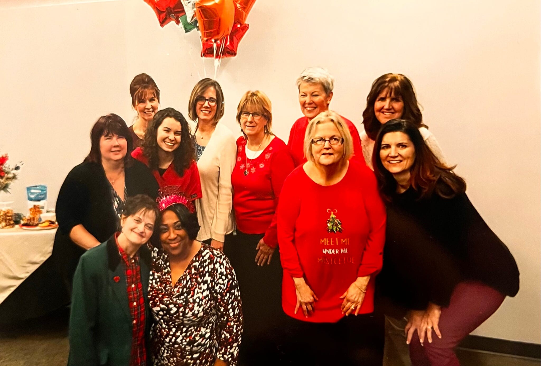 Group of women posing together at a holiday party, several in red outfits, smiling with balloons in the background.
