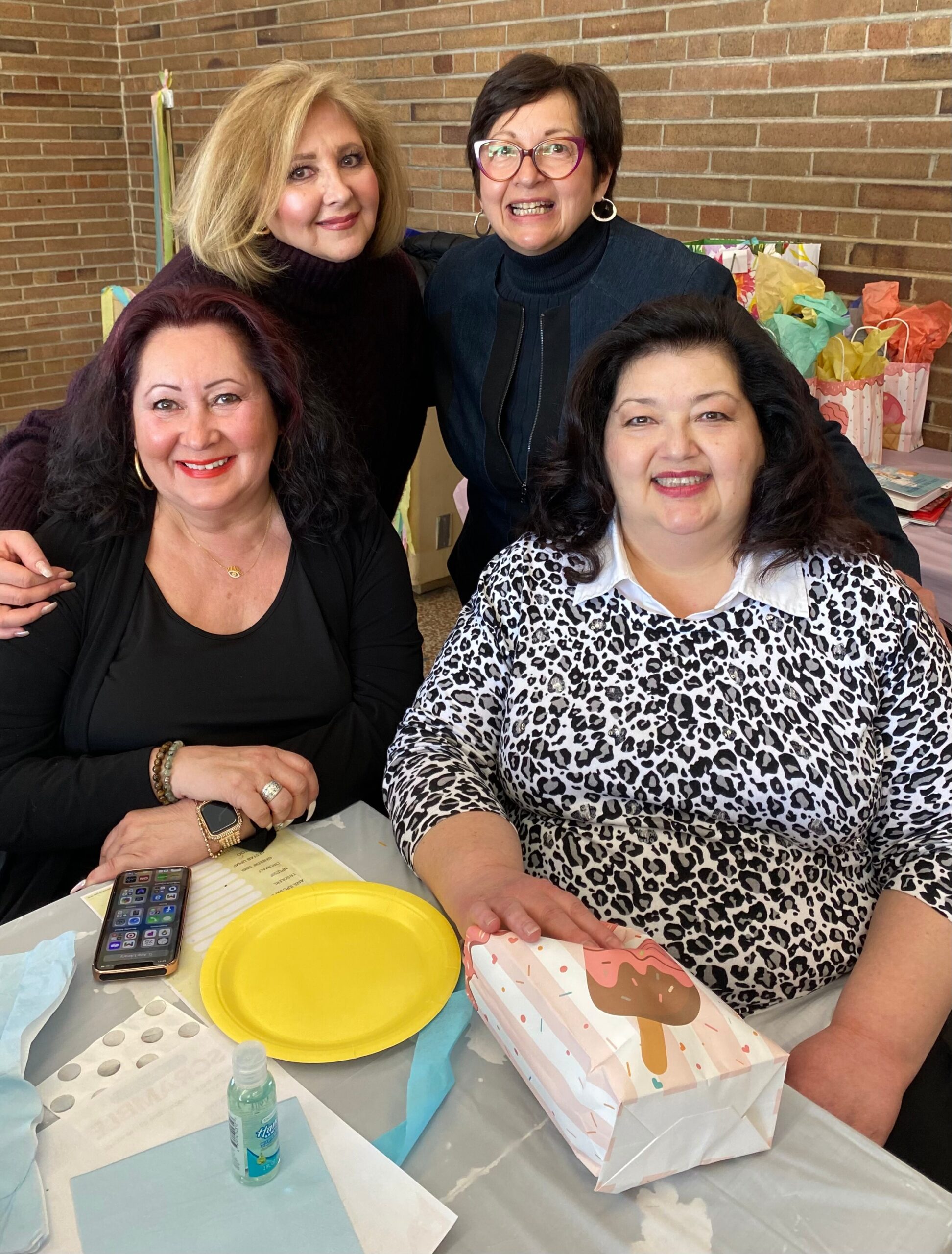 Four women smiling at a party around a table with a yellow plate and colorful gift bags in the background.