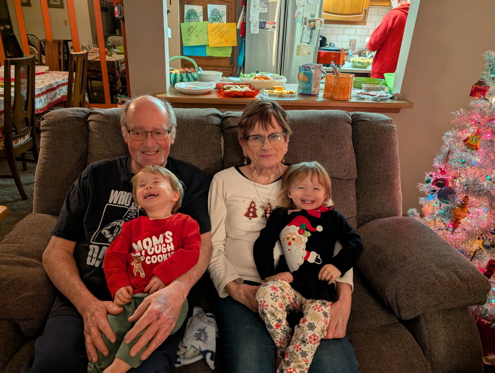 Grandfather, grandmother, and two young children sit together smiling on a brown couch in a festive living room.
