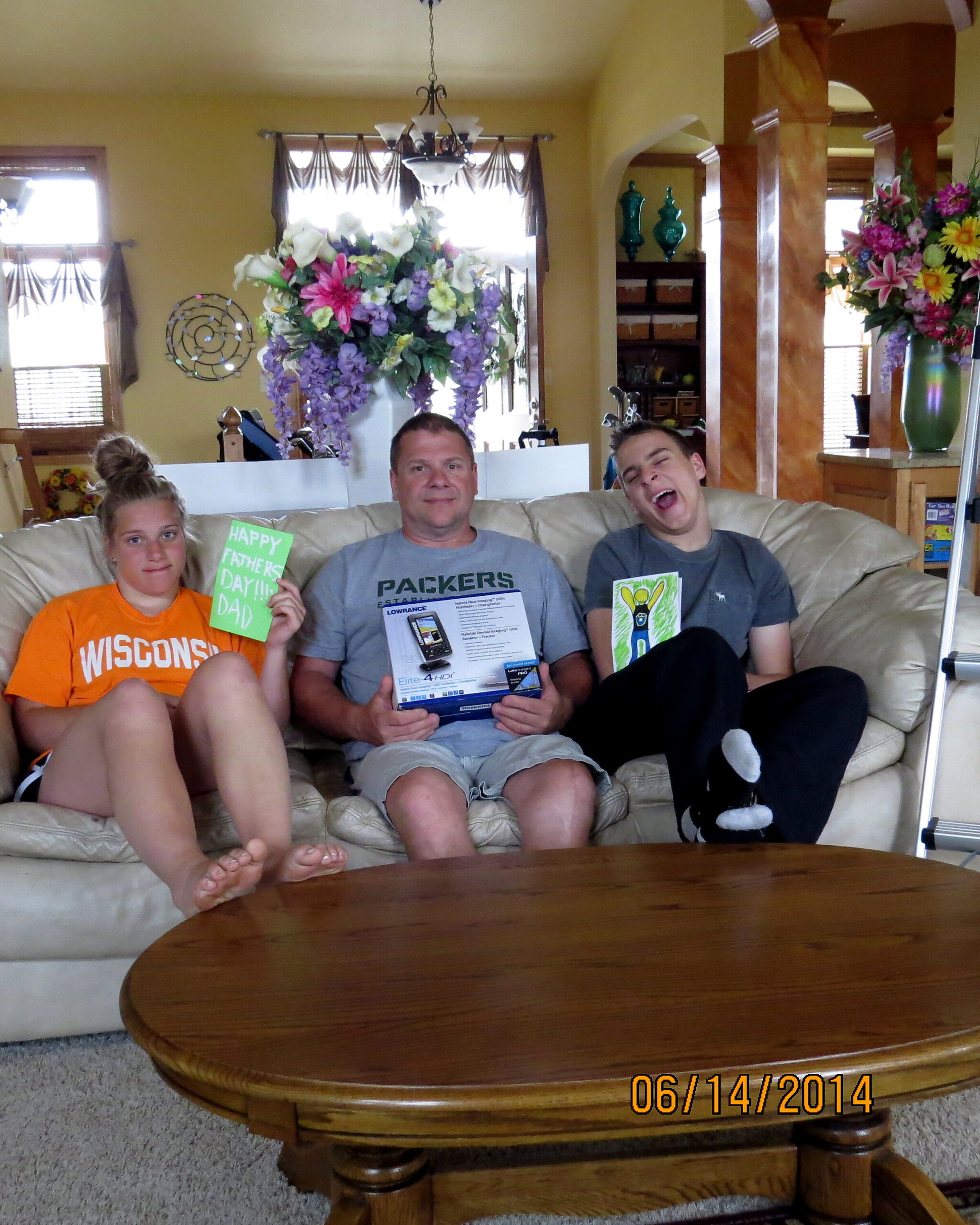A family sits on a beige sofa in a sunlit living room, holding greeting cards on Father’s Day, with a wooden coffee table in front.