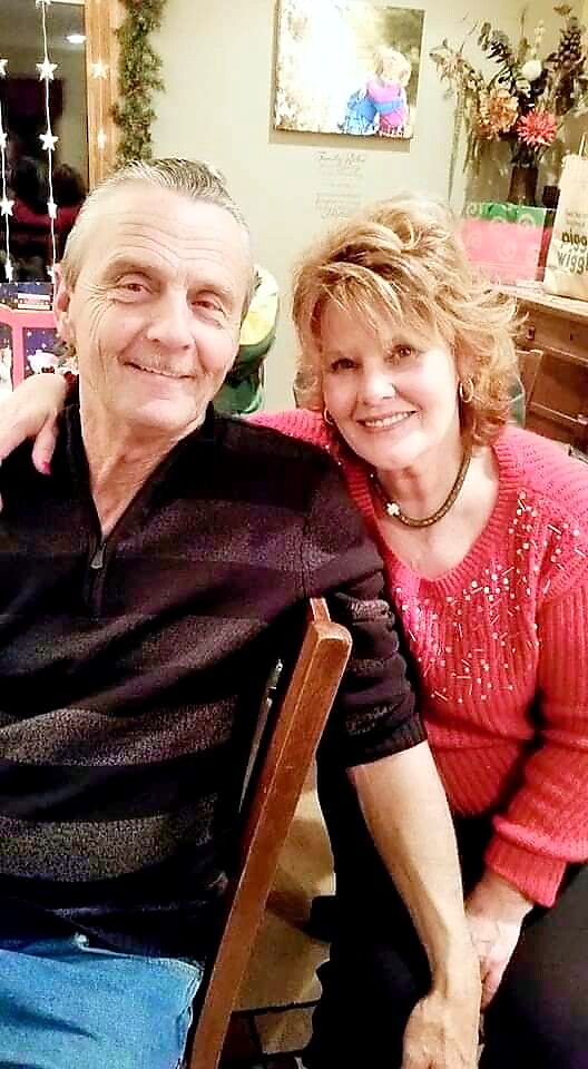 Smiling older couple posing together with the woman in a red sweater and their arms around each other in a cozy, decorated room.