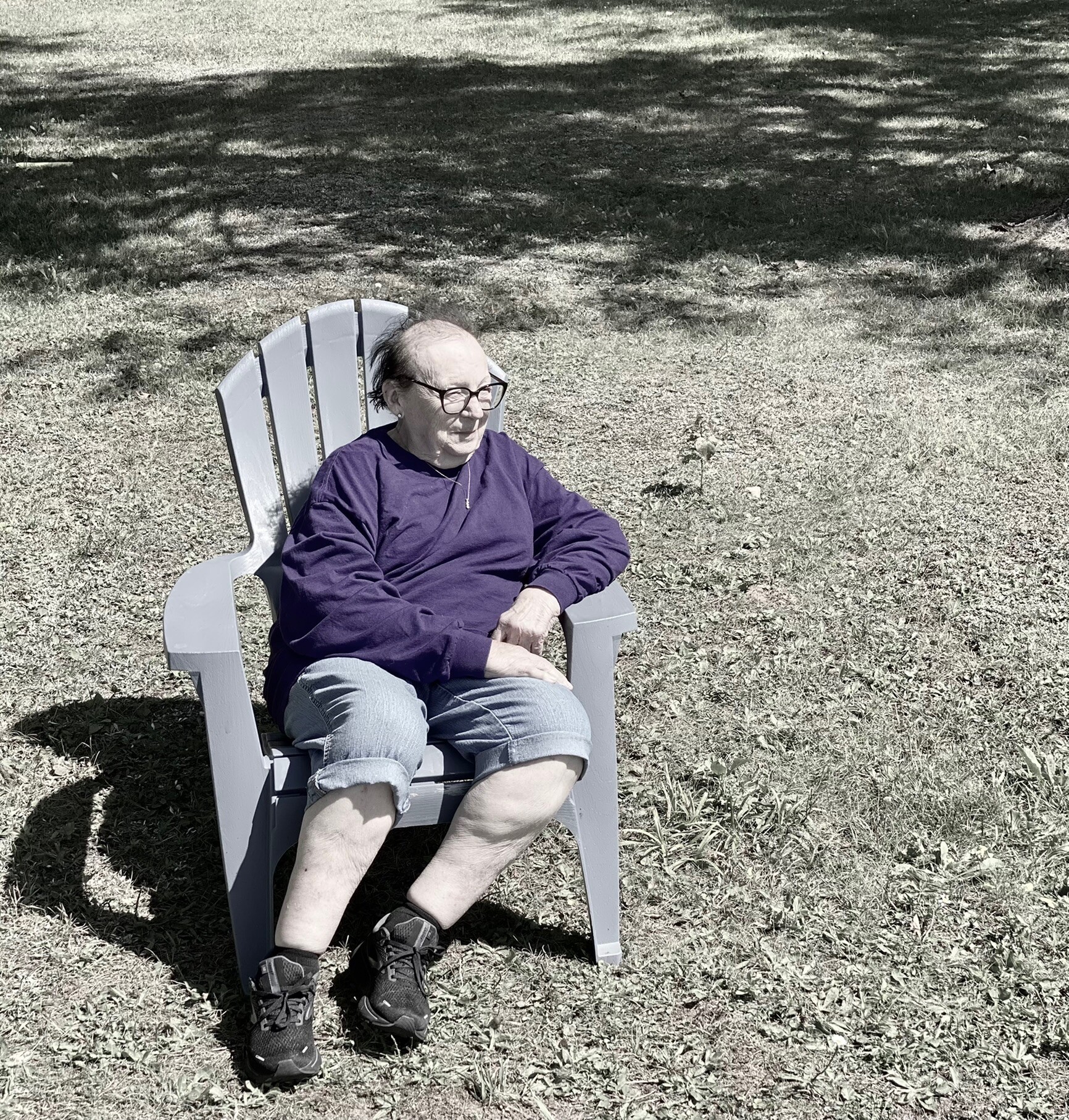 Older person sitting in a gray plastic lawn chair on a grassy yard, wearing a purple long-sleeve shirt and jeans, smiling outdoors.