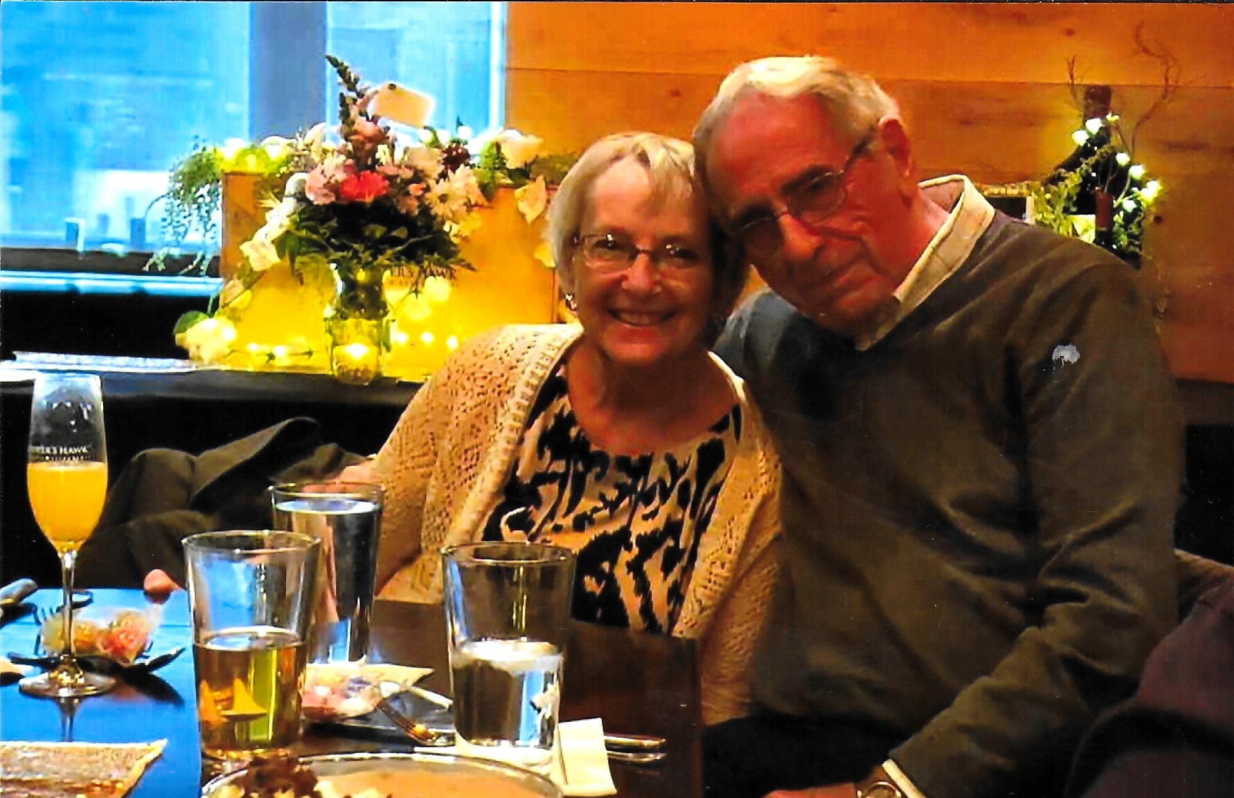 Old couple smiling and leaning together at a dining table during a celebration, with drinks and desserts in front of them and floral decor in the background.