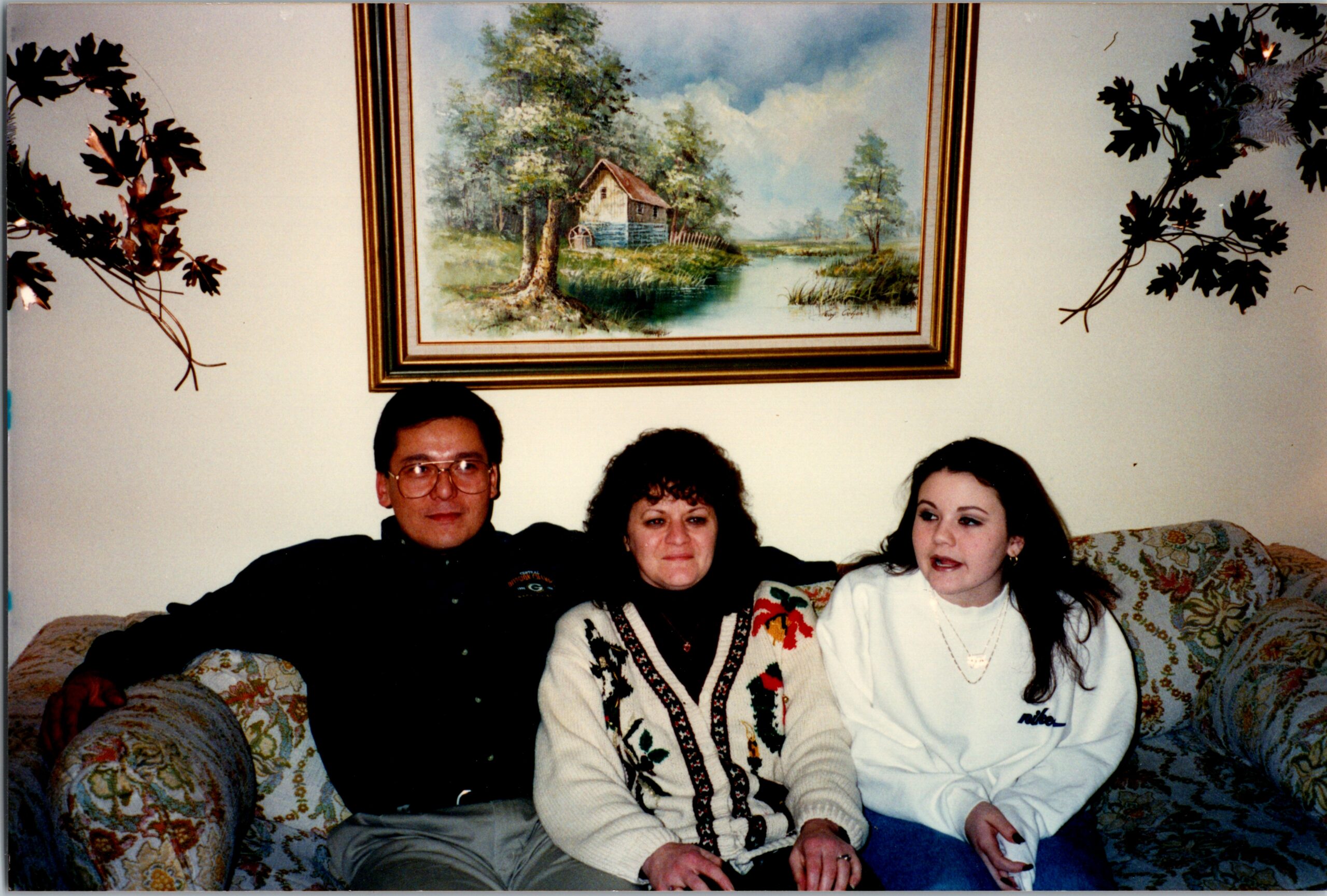 Three people sit together on a floral sofa in a living room; a landscape painting hangs on the wall behind them.