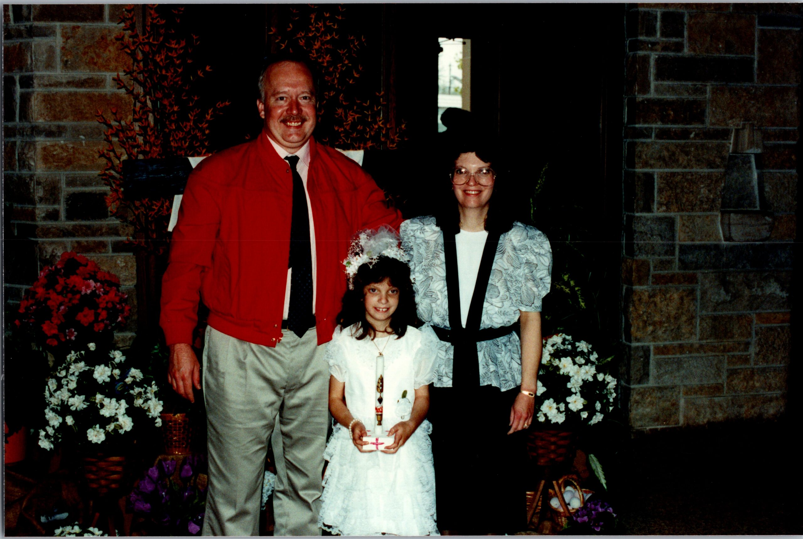 Family posing for a photo outside a brick building; man in a red jacket, woman in a patterned blazer, and two young girls in white dresses with a candle centerpiece behind them.
