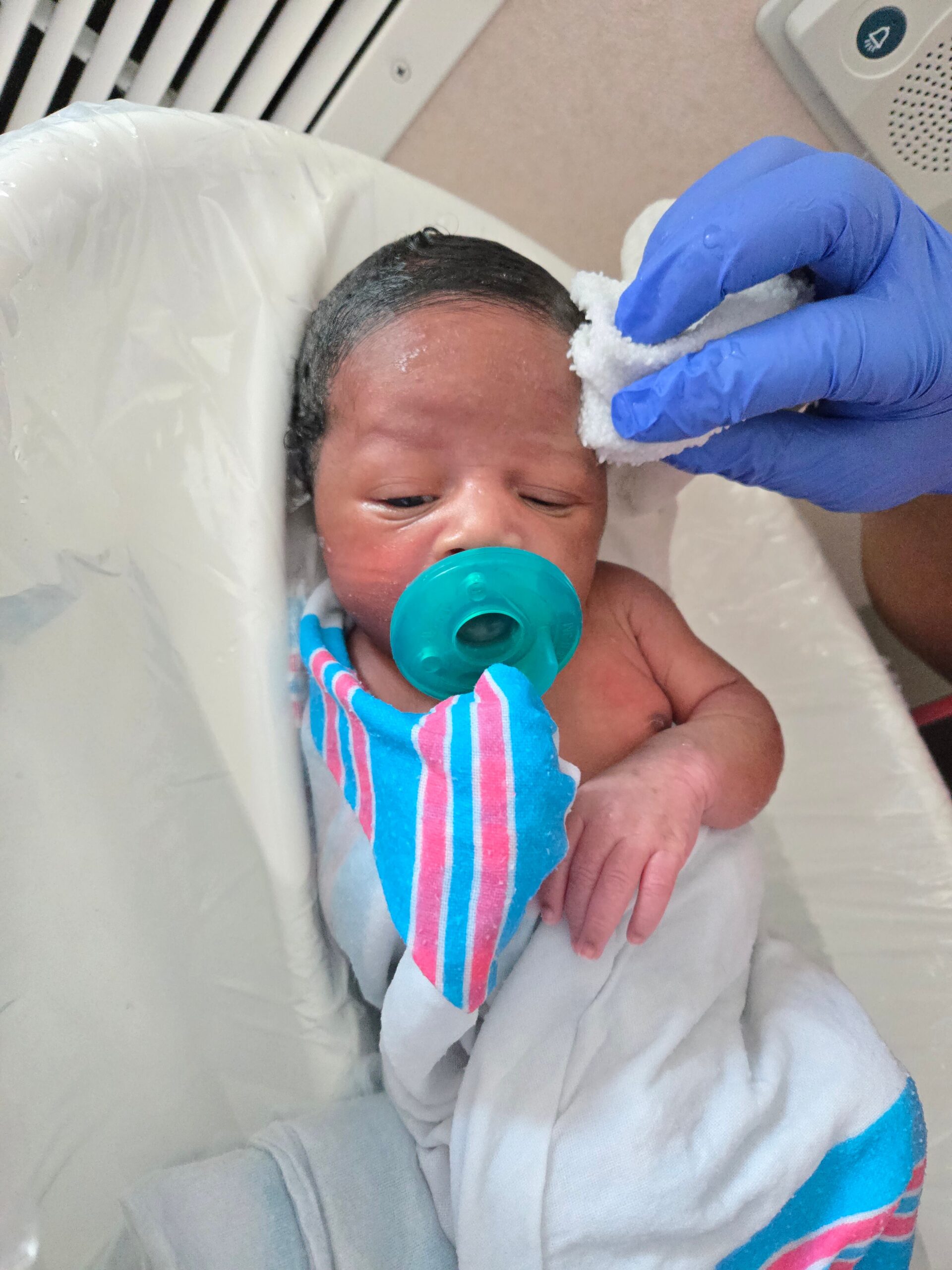 Medical staff cleans a sleeping baby's forehead with a gauze pad while the infant wears a teal pacifier and striped blanket.