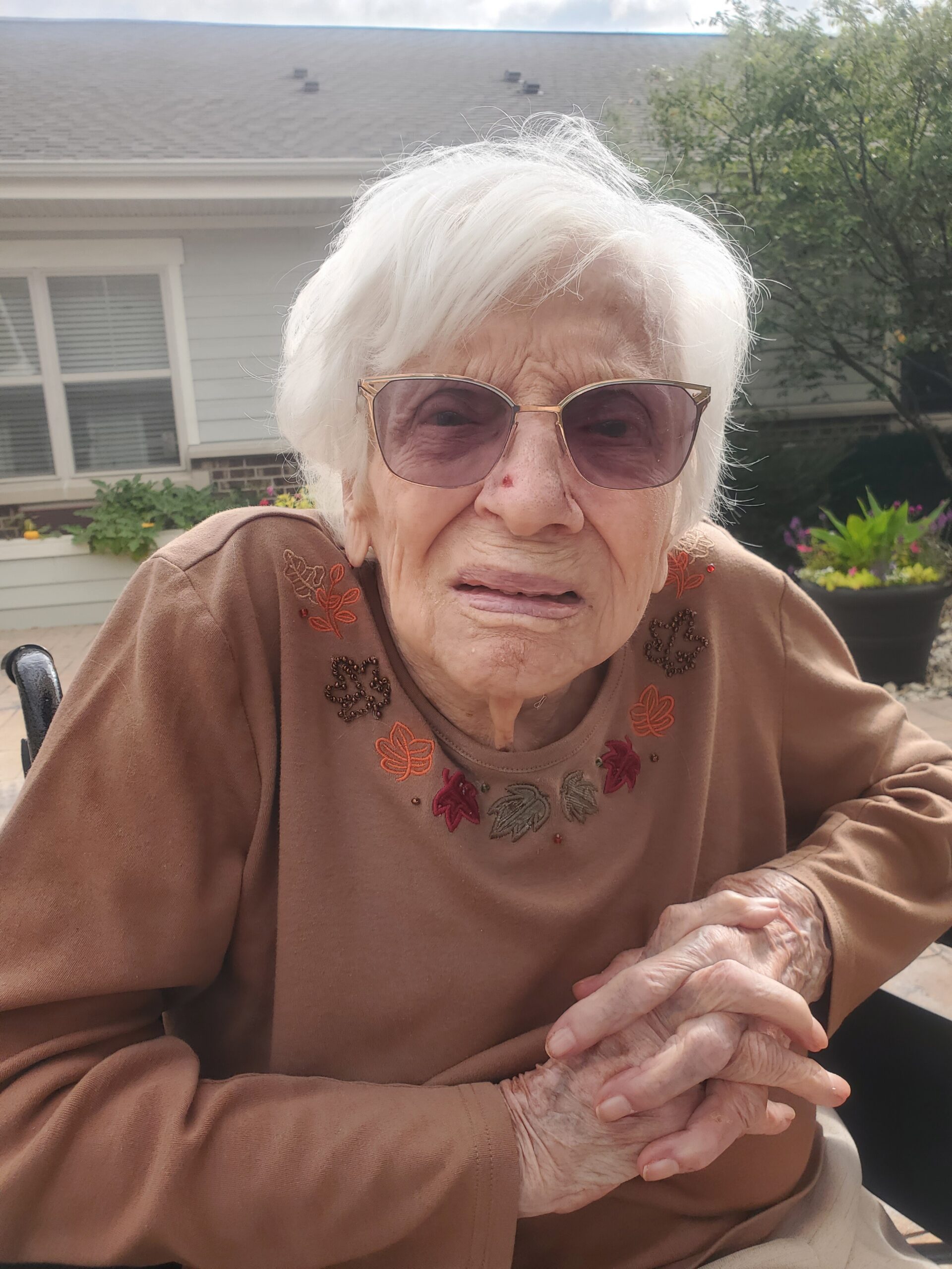 Elderly woman with white hair and pink-tinted glasses sits outdoors, hands clasped, wearing a brown embroidered top.