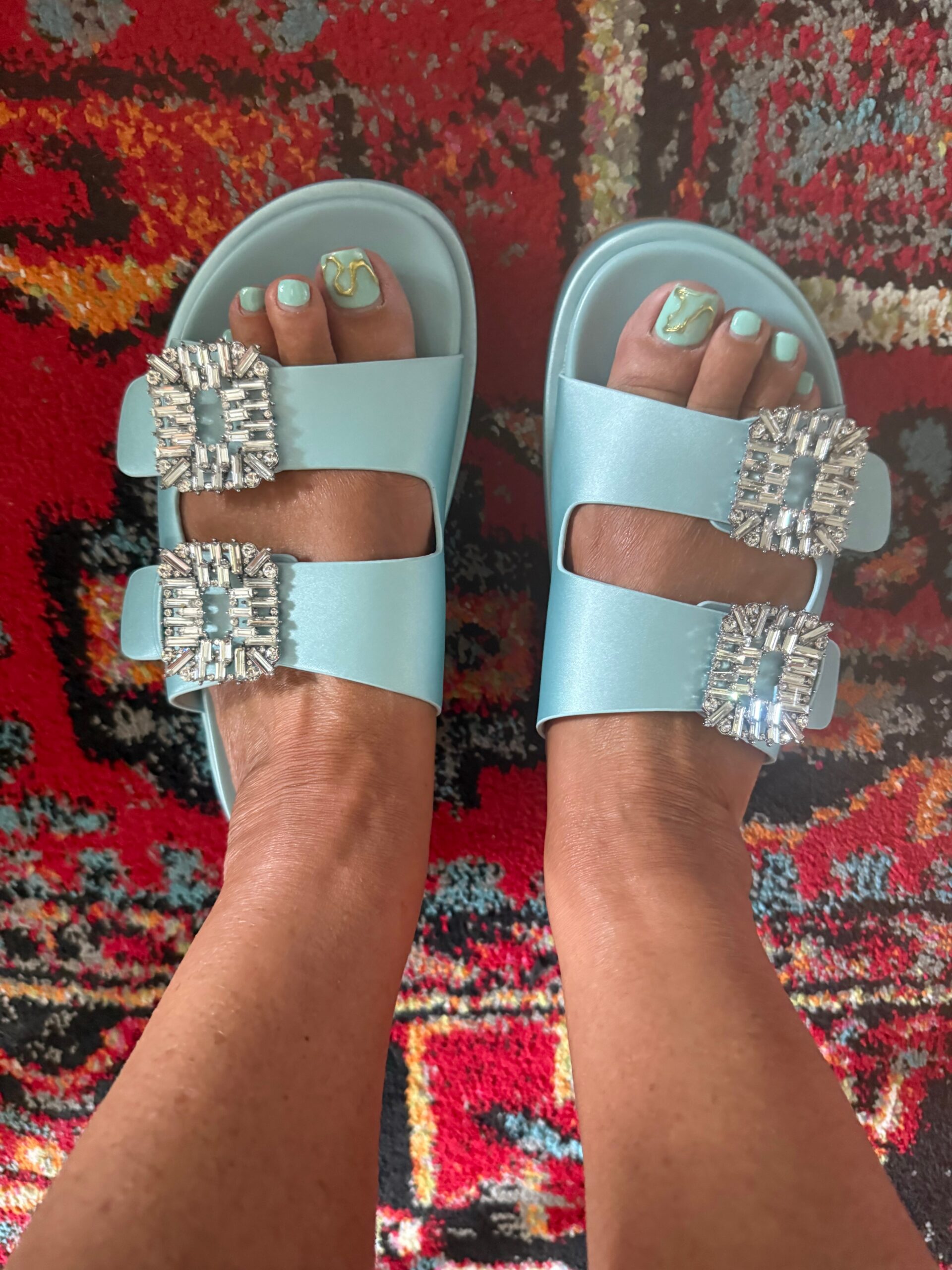 Mint-green painted toenails in light blue slide sandals with large jeweled square buckles, standing on a colorful patterned rug