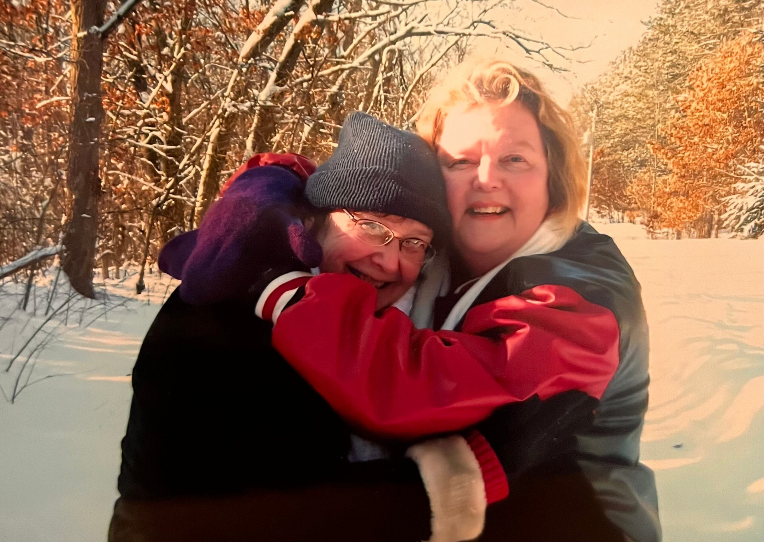 Two people hugging outdoors in a snowy forest, both wearing winter jackets and smiling warmly at the camera.