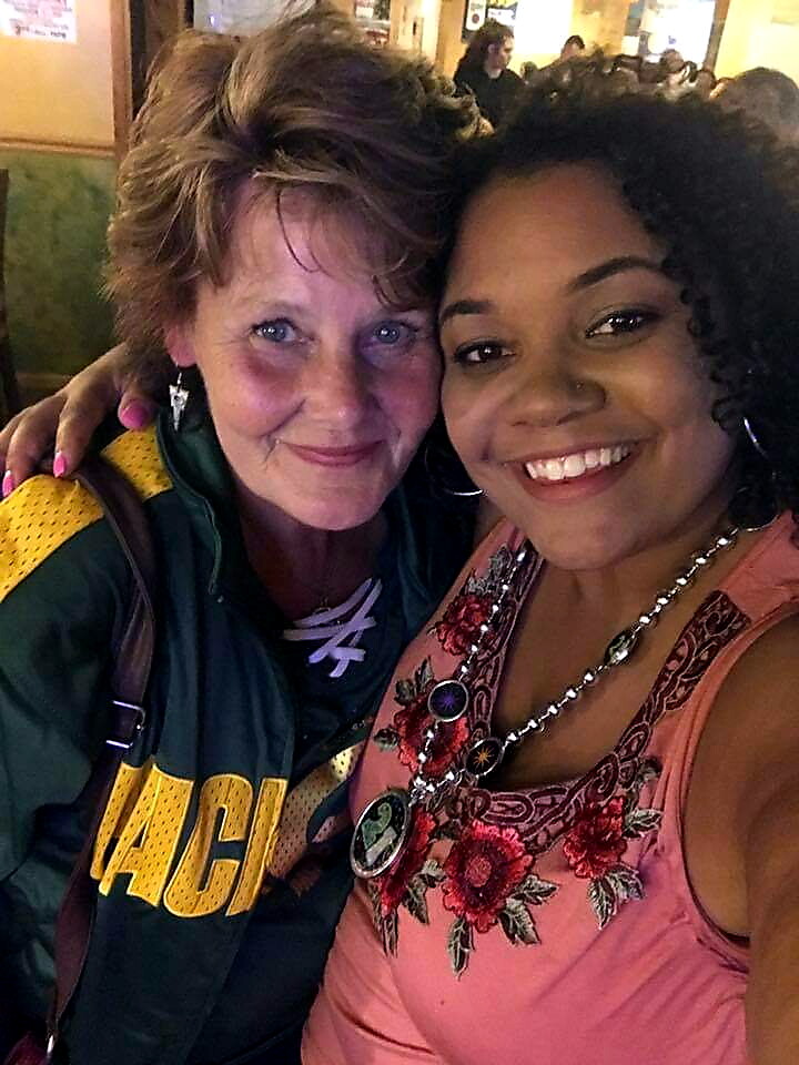 Two smiling women pose for a close selfie at a busy indoor venue; one in a green-yellow jacket with 'PACK' lettering, the other in a pink floral top with a statement necklace.
