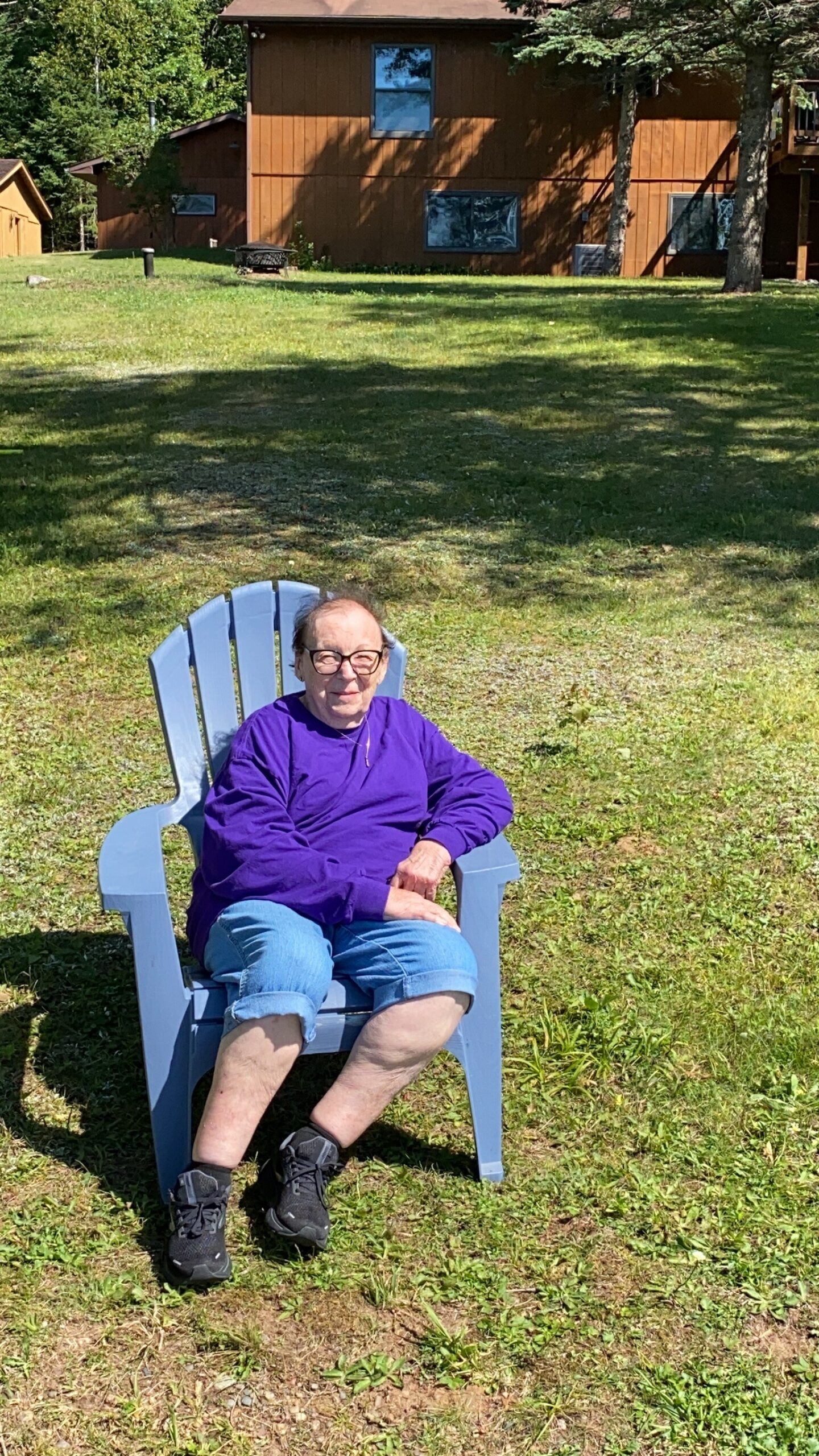 Elderly person in a purple top sitting in a blue lawn chair on a grassy yard with a brown house in the background.