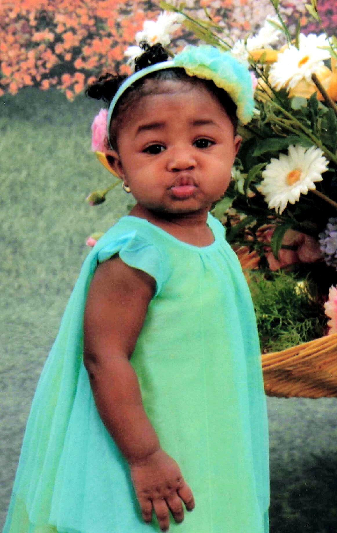 Toddler girl in a turquoise dress with a pastel flower headband outdoors among flowers, facing the camera and pursing her lips.
