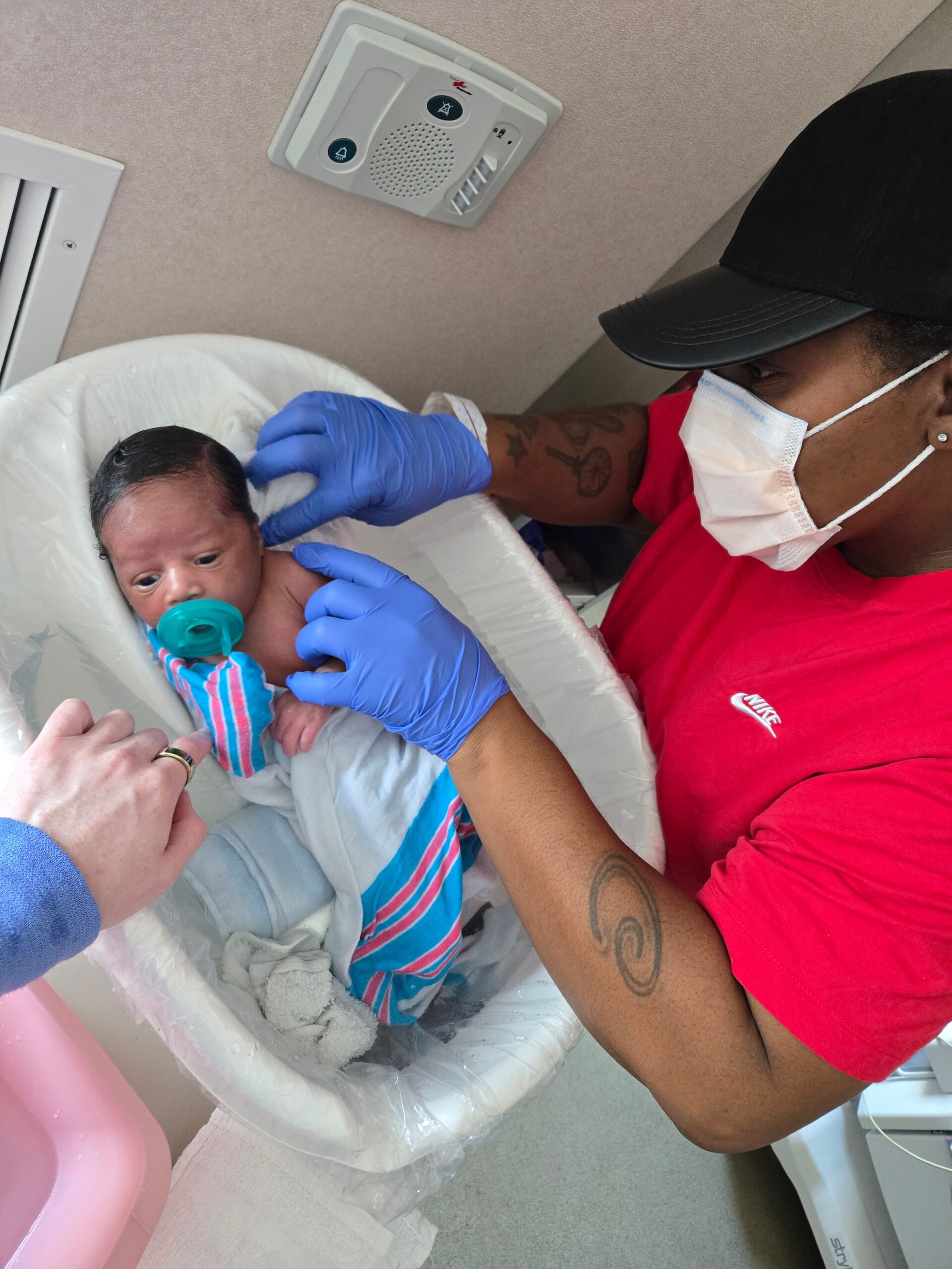Caregiver wearing a mask and blue gloves tends to a newborn baby lying in a bassinet, baby with a teal pacifier wrapped in a striped blanket.